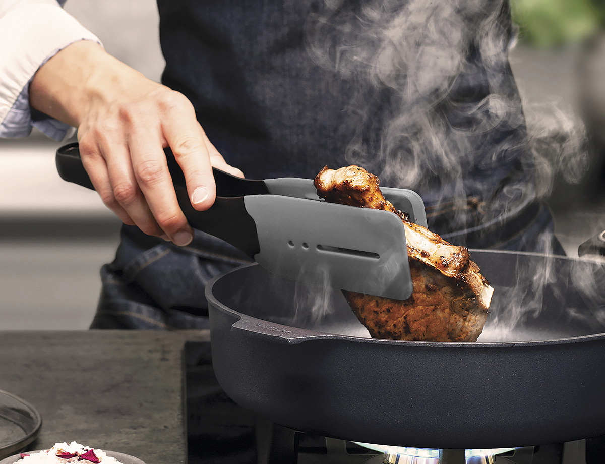 Ein dampfendes, angebratenes Stück Fleisch wird mithilfe der grauen Cock it Küchenzange in der heißen Pfanne gewendet.