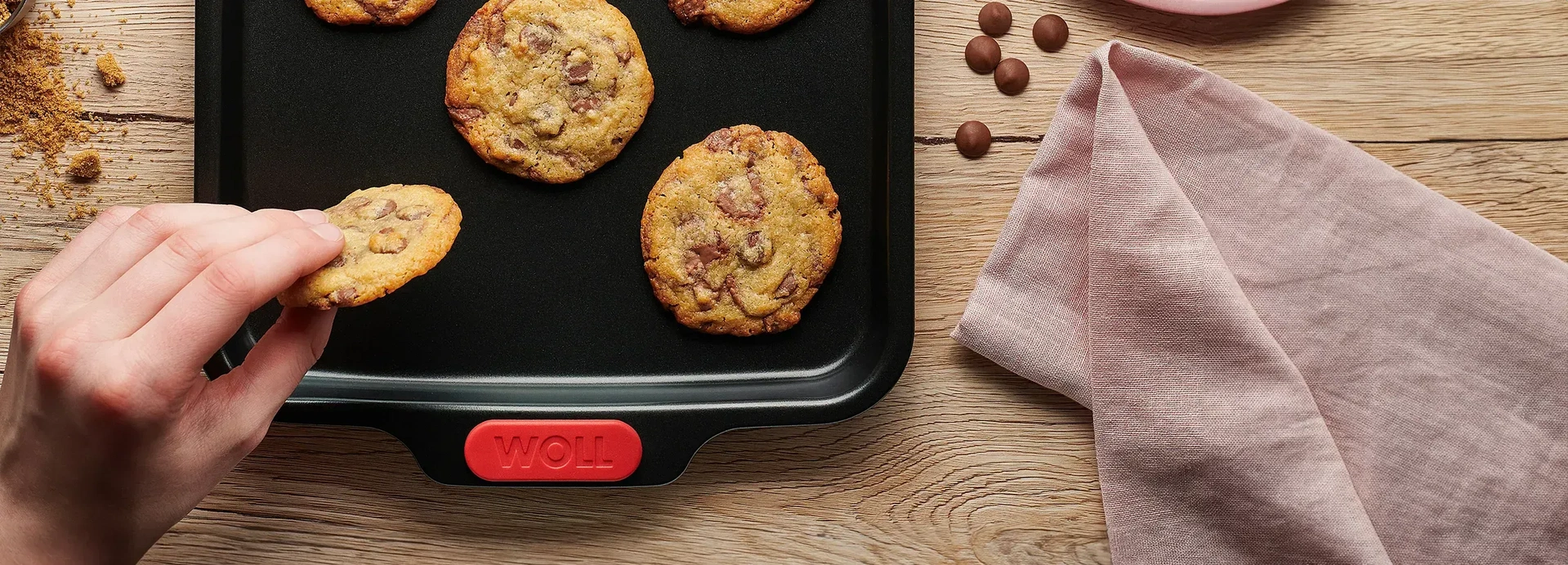 Auf einem Backblech der Serie Let`s Bake! liegen frischgebackende Cookies, als Deko liegen Schokodrops, eine Servierte auf dem Tisch.