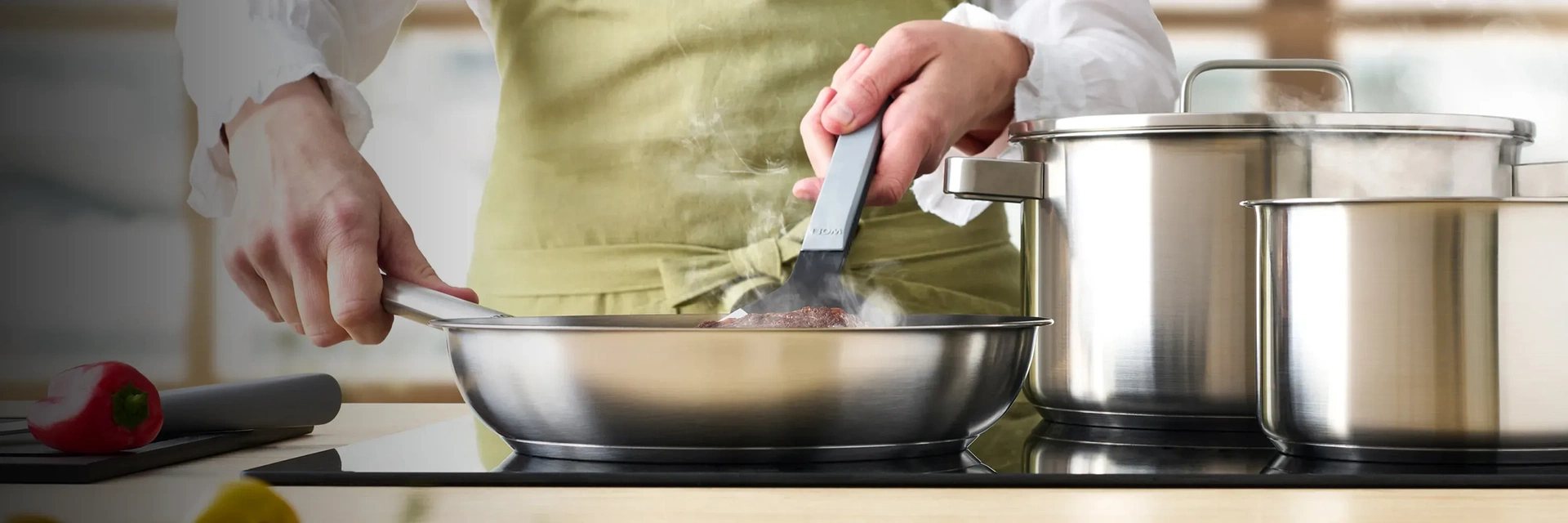 In einer runden, heißen Pfanne der Serie „Steel” wird Fleisch angebraten und mit einem Pfannenwender gewendet. Daneben stehen zwei Töpfe in unterschiedlichen Größen. Auf der Arbeitsplatte liegt ein Schneidebrett mit einer Paprika darauf, daneben liegt ein