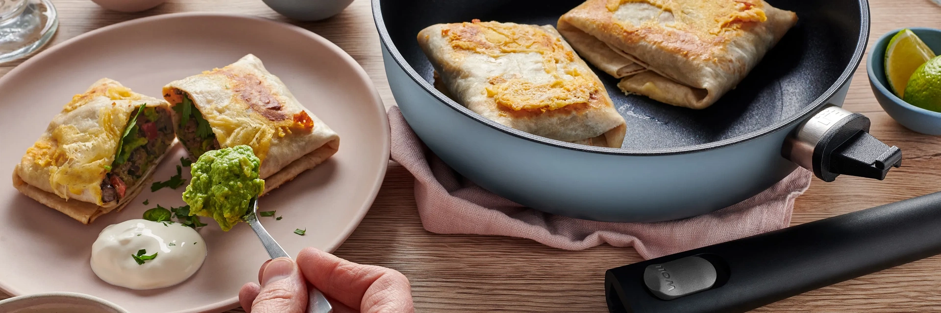 In einer runden Ecolour-Pfanne mit abgenommenem Stiel befinden sich frisch angebratene Burritos. Ein Burrito ist in der Mitte geteilt und liegt auf einem Teller. Er wird mit zwei Dips serviert.