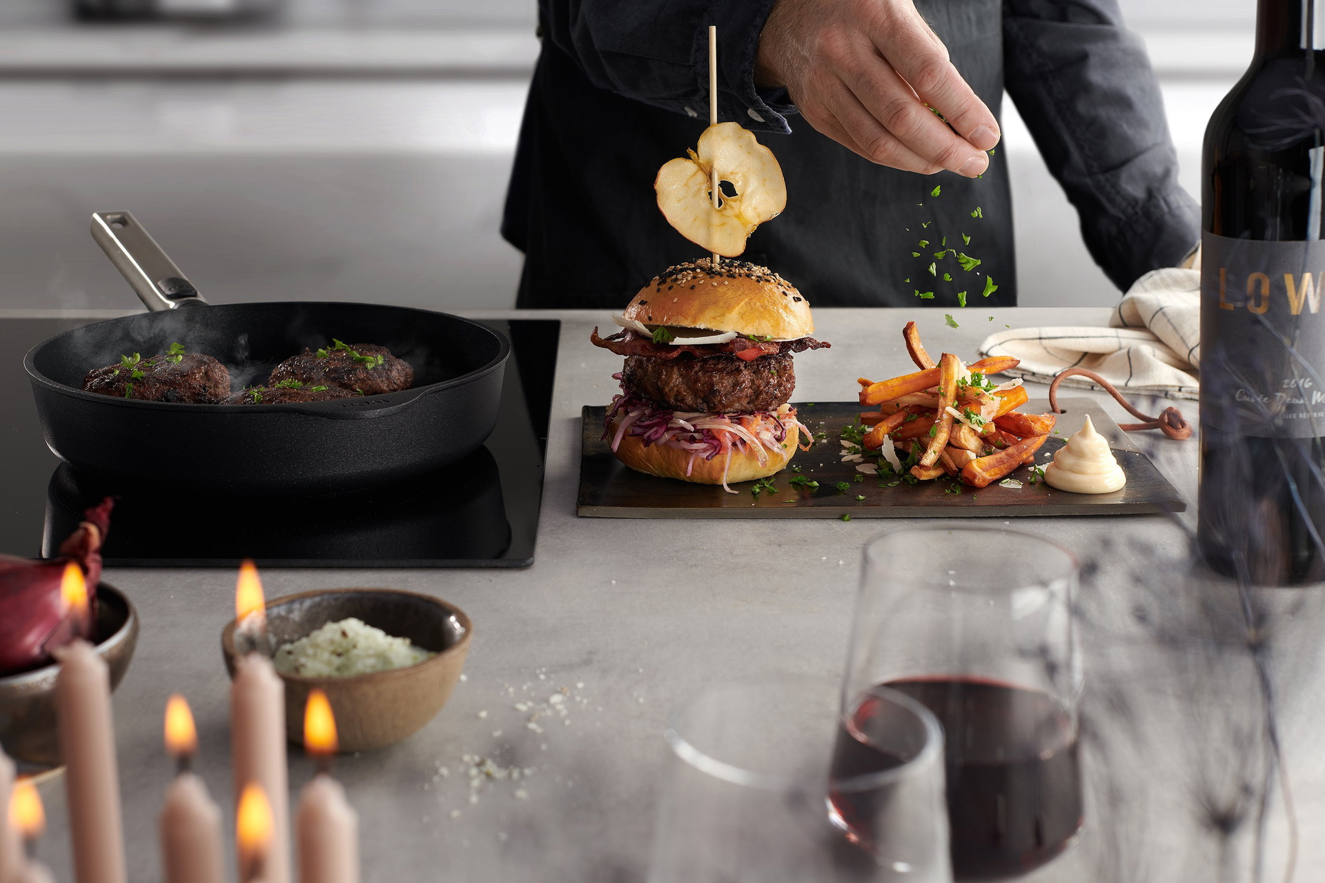 In einer runden Logic XR Pfanne werden Burgerpattys angebraten. Auf einem Brett steht ein angerichteter Burger.