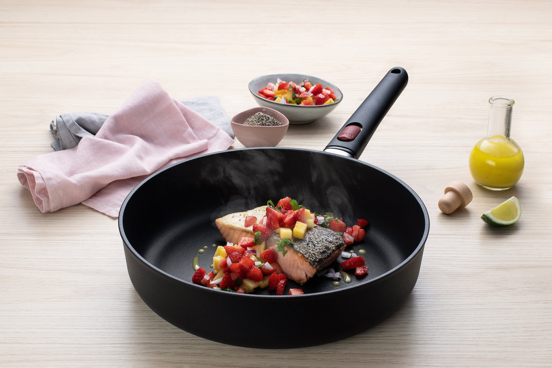 In einer heißen, runden Eco-Lite-QXR-Pfanne mit abnehmbarem Stiel befindet sich frisch angebratener Lachs mit Mango-Salsa. Daneben steht eine Schüssel mit Mango-Salsa, Gewürz, zwei Geschirrtücher und frisch gepresstem Zitronensaft.