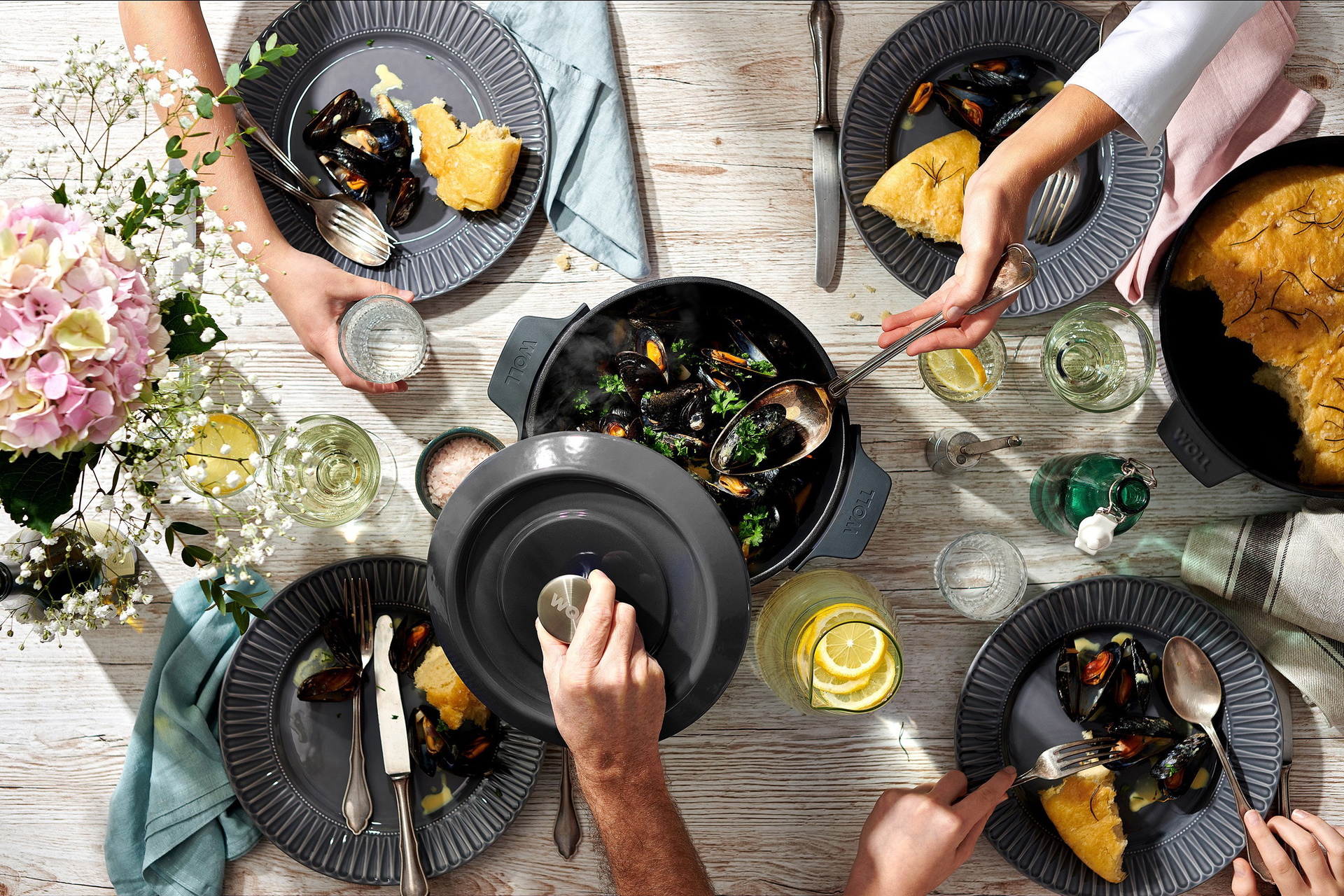 Auf dem Tisch steht eine runde, graue Iron-Kasserolle mit zubereiteten Muscheln. Der Deckel wird hochgehoben.