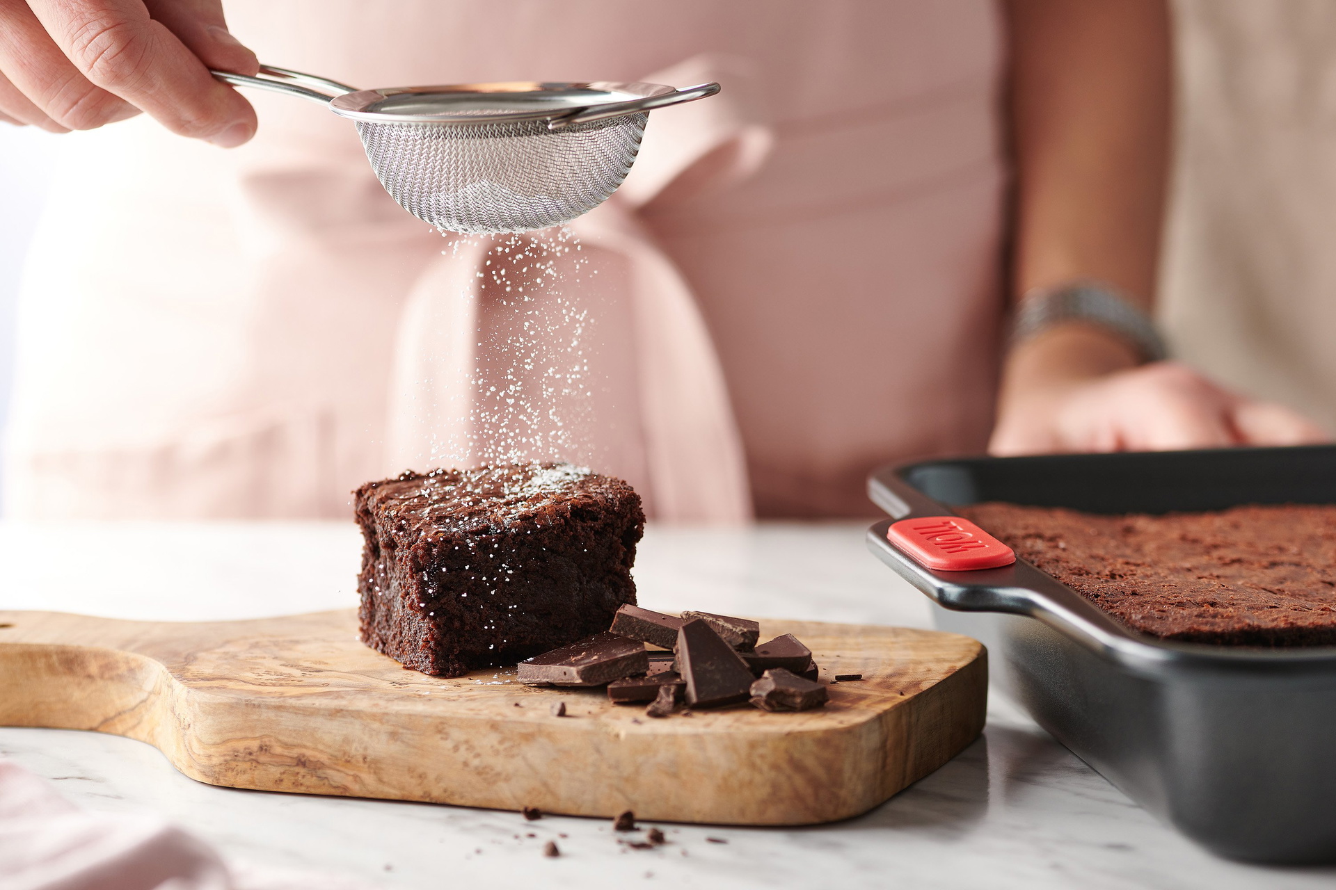 Auf einem Holzbrett liegt ein frisch gebackenes Stück Brownie, das mit Puderzucker bestäubt ist. Schokoladenstückchen auf dem Holzbrett dienen als Dekoration. Daneben steht die rechteckige Backform der Let's Bake!-Serie, die mit weiterem Brownieteig gefül
