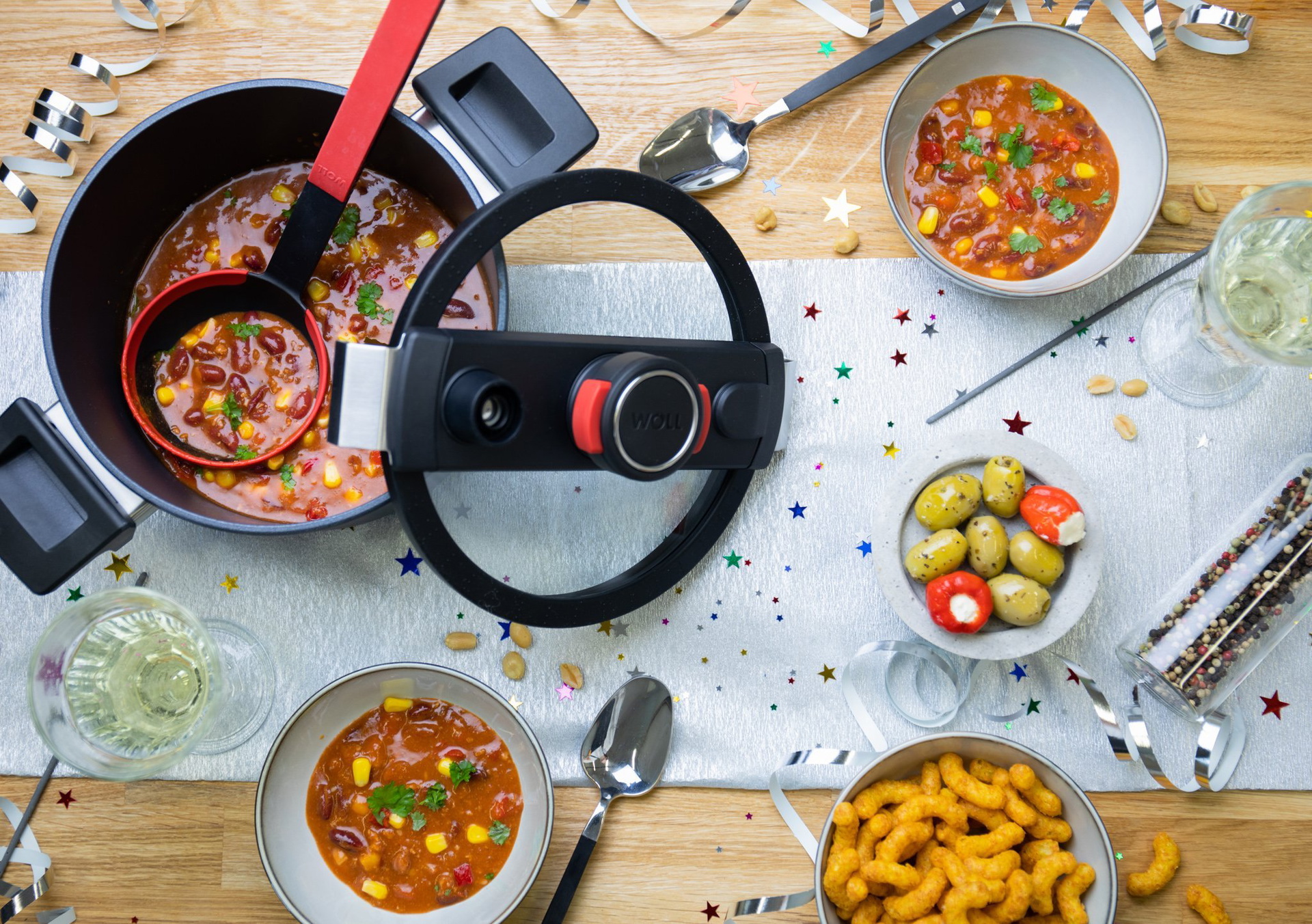 Auf einem Tisch steht ein runder Niederdrucktopf der Serie „Diamond Active Lite Gusstopf”, dessen Deckel schräg am Topf angelehnt ist. In ihm befindet sich frisches, gekochtes Chili con Carne. Drumherum stehen Schüsseln mit Chili con Carne, gefüllte Olive