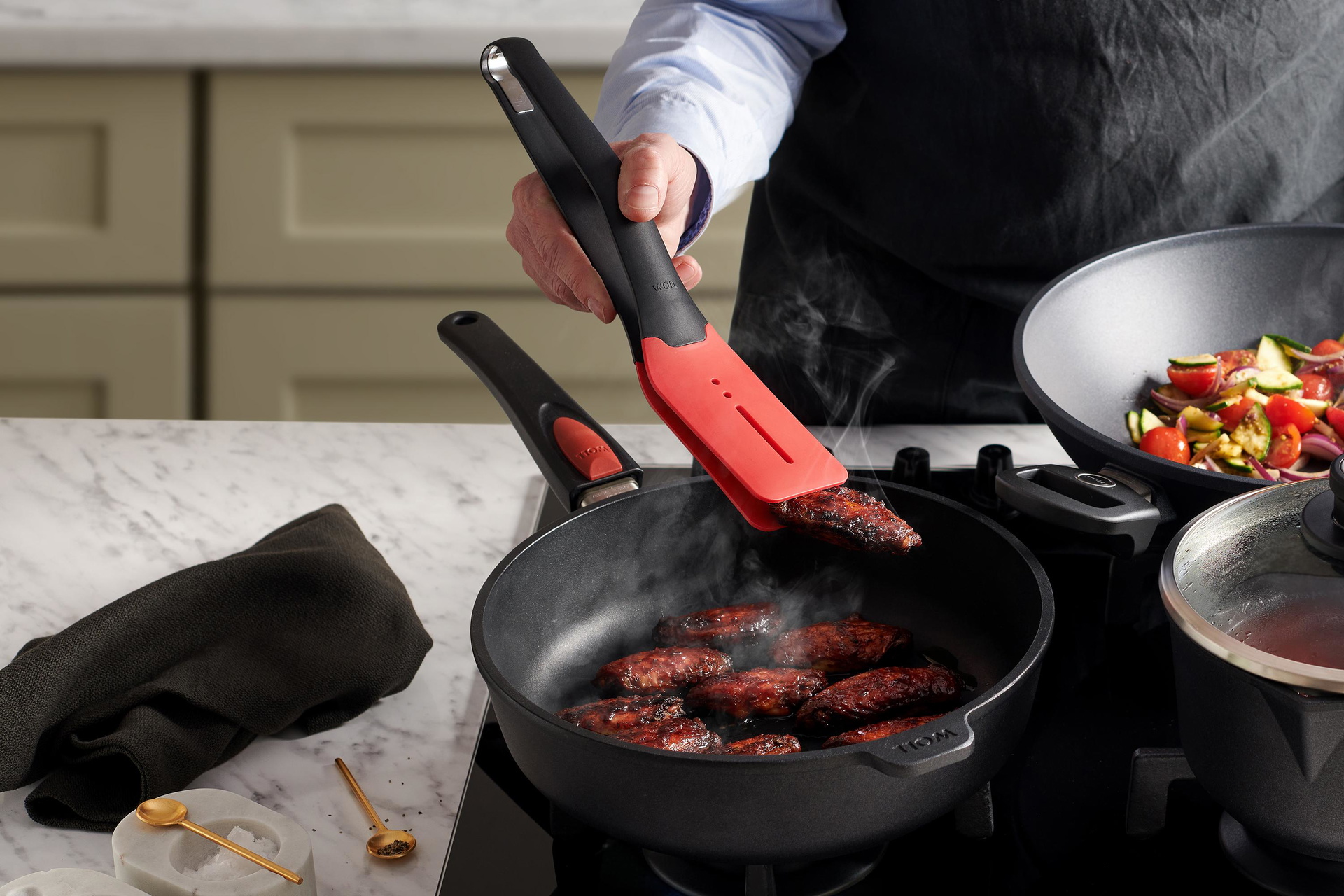 In einer runden Titan Best Pfanne werden Fleischstücke angebraten und mit einer Cock it Küchenzange gewendet.