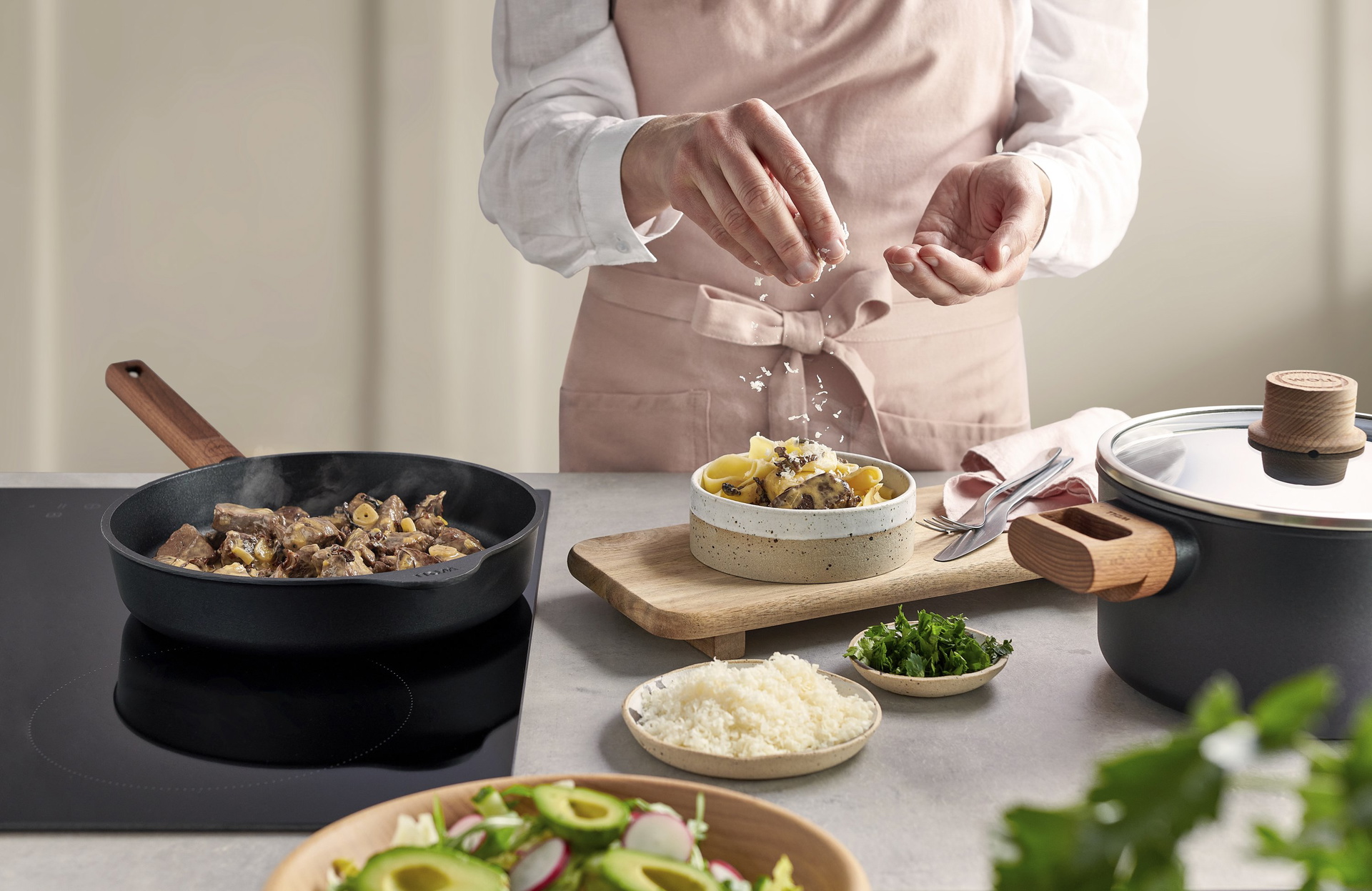 Auf dem Herd steht eine runde, heiße Eco-Logic-QXR-Pfanne mit festem Holzstiel, in der Rinderfiletstreifen mit Pfifferlingen angebraten werden. Daneben wird eine Portion Rinderfilet mit Pappardelle in einer Schüssel angerichtet. Drumherum liegen Besteck,