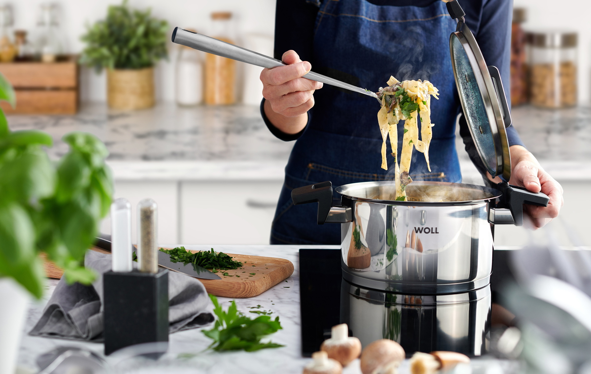 Im runden Edelstahltopf der Serie „Concept” befinden sich frisch zubereitete Cremige Pilz-Tagliatelle. Der Deckel wurde abgenommen und in die dafür vorgesehene Halterung am Griff gesteckt. Daneben liegt ein Schneidebrett mit frischen Kräutern und einem Me