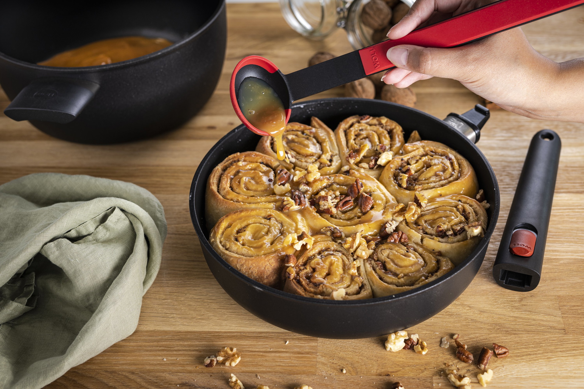 1724ELI_124ELI_Zimtschnecken_3000x2000px In einer runden Eco Lite Pfanne befinden sich Zimtschnecken, diese werden mit einem roten Cock it Saucenlöffel mit Karamellsoße übergossen.