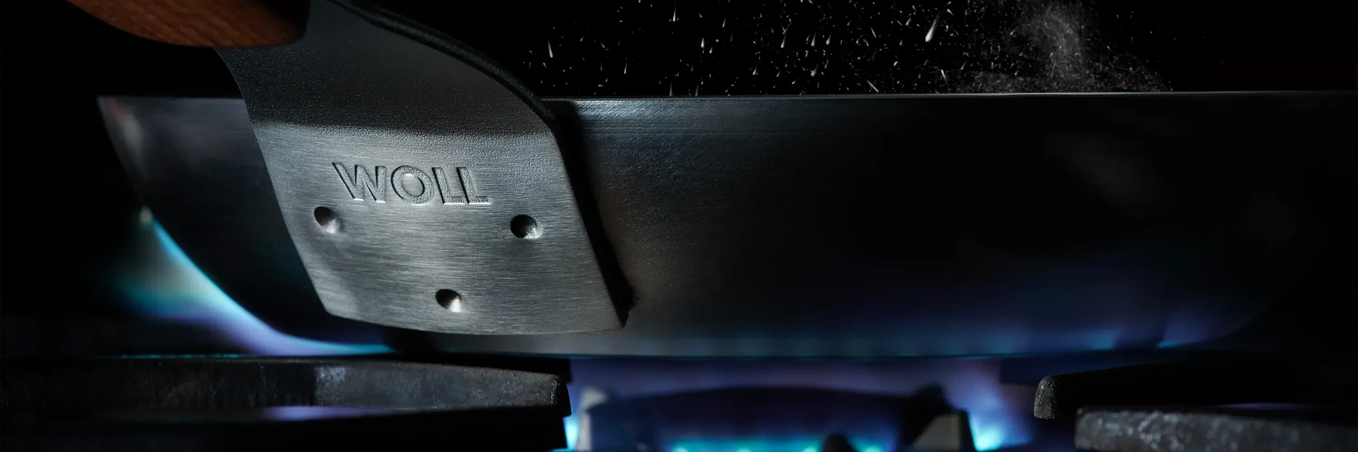 In einer Nahaufnahme sind die Befestigungspunkte des Griffs an der Carbon-Steel-Pfanne zu erkennen. Die Pfanne steht auf einem Gasherd, darunter brennen blaue Flammen.