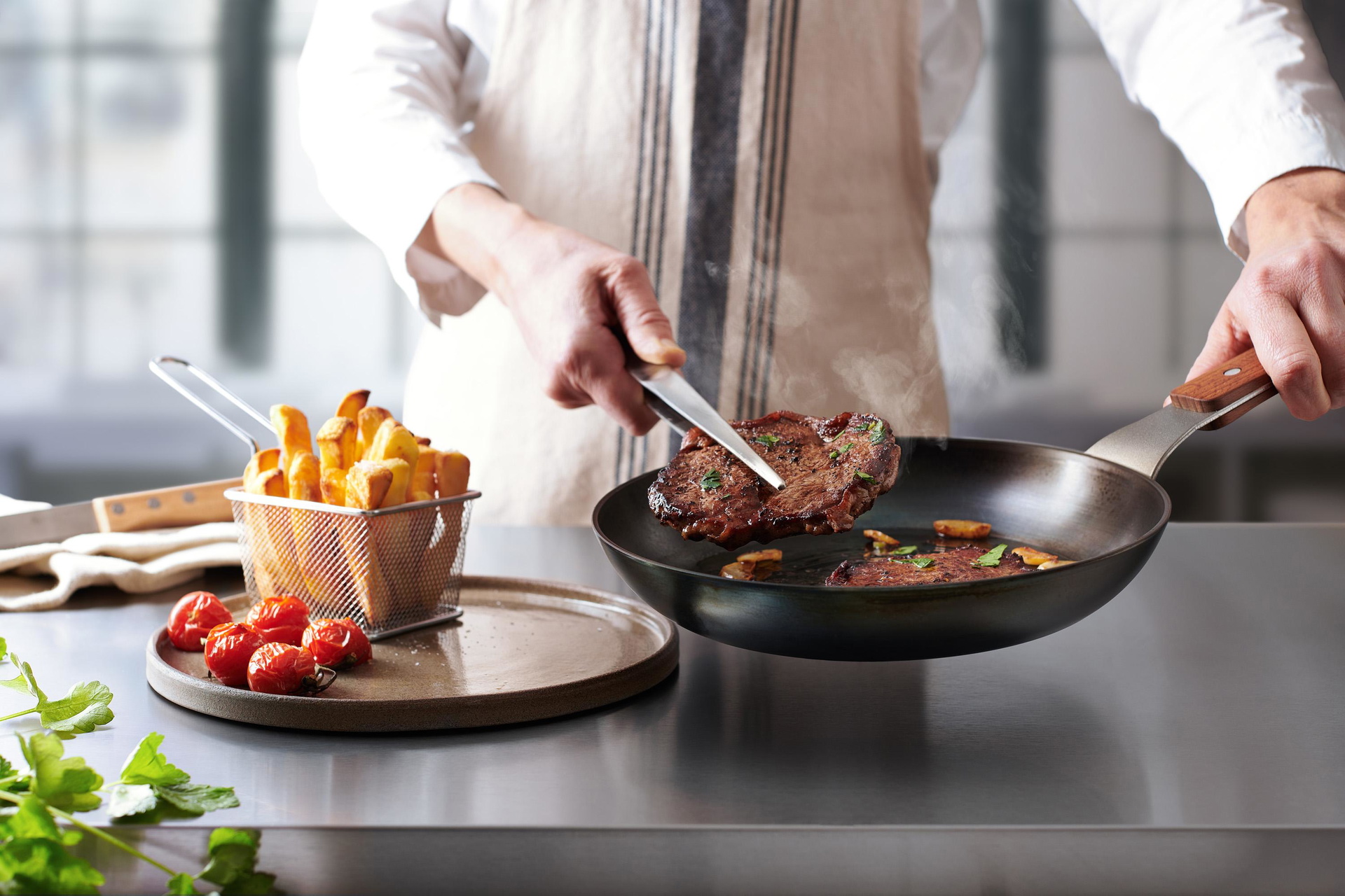 Carbon Steel Flachpfanne In einer runden Carbon Steel Pfanne befinden sich angebratene Steaks. Ein Steak wird auf einem Teller platziert.