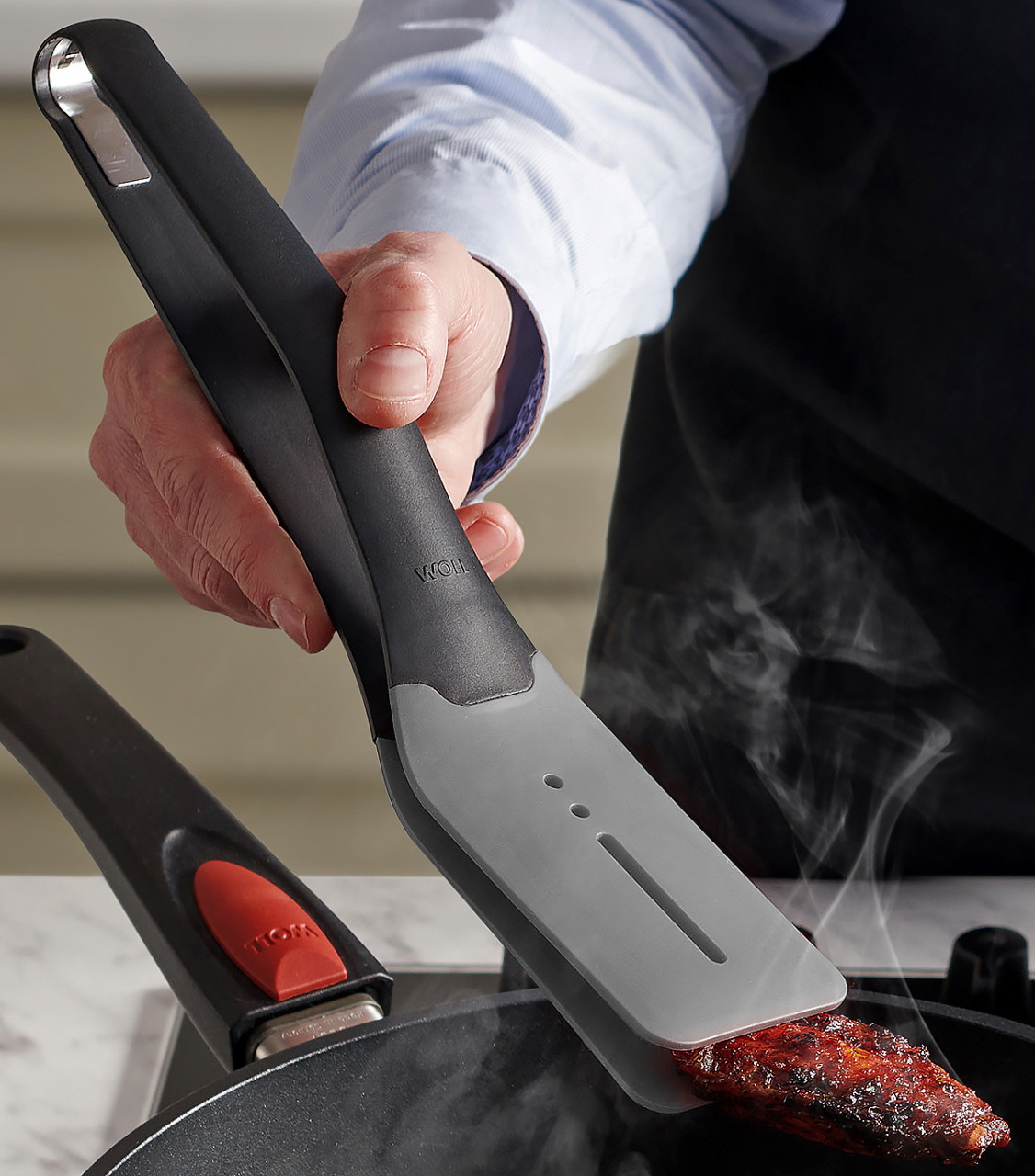 Ein dampfendes, angebratenes Stück Fleisch wird mithilfe der grauen Cock it Küchenzange in der heißen Pfanne gewendet.