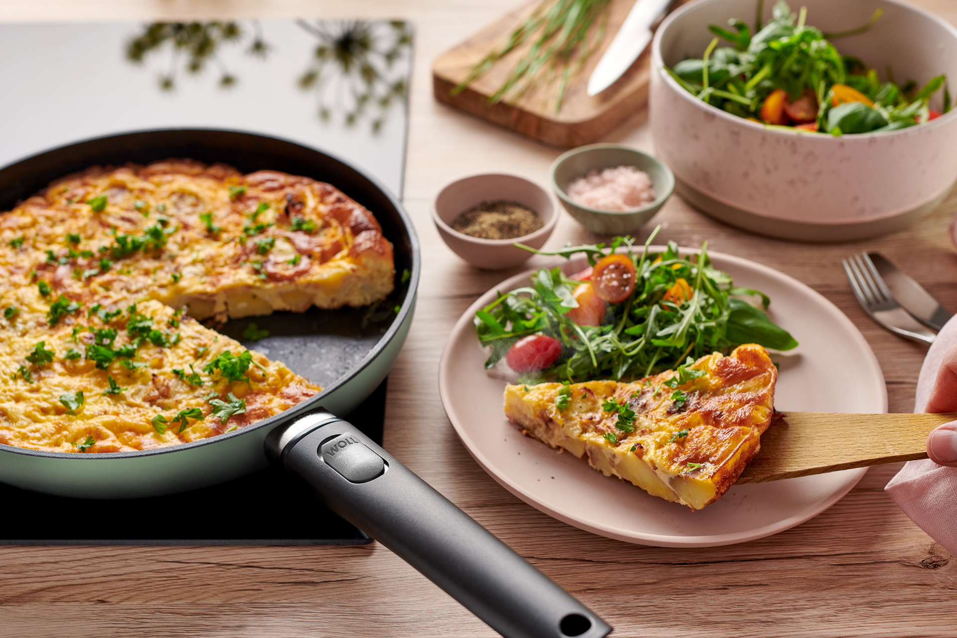 In einer runden, jadegrünen Ecolour Pfanne befindet sich eine frische spanische Tortilla. Ein Stück davon liegt dem Teller.