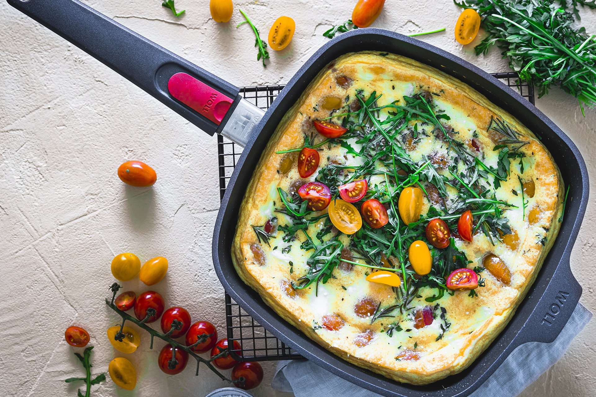 In einer viereckigen Diamond-Lite-Pfanne mit abnehmbarem Stiel befindet sich ein Clafoutis mit bunten Kirschtomaten und Mozzarella. Drumherum liegen weitere Kirschtomaten, Kräuter und Salz.