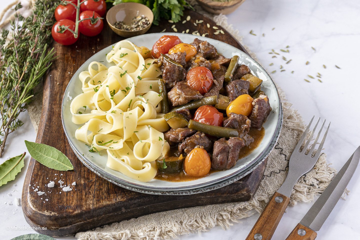 Auf einem Holzbrett steht ein Teller mit mediterranem Lammragout und Bandnudeln.