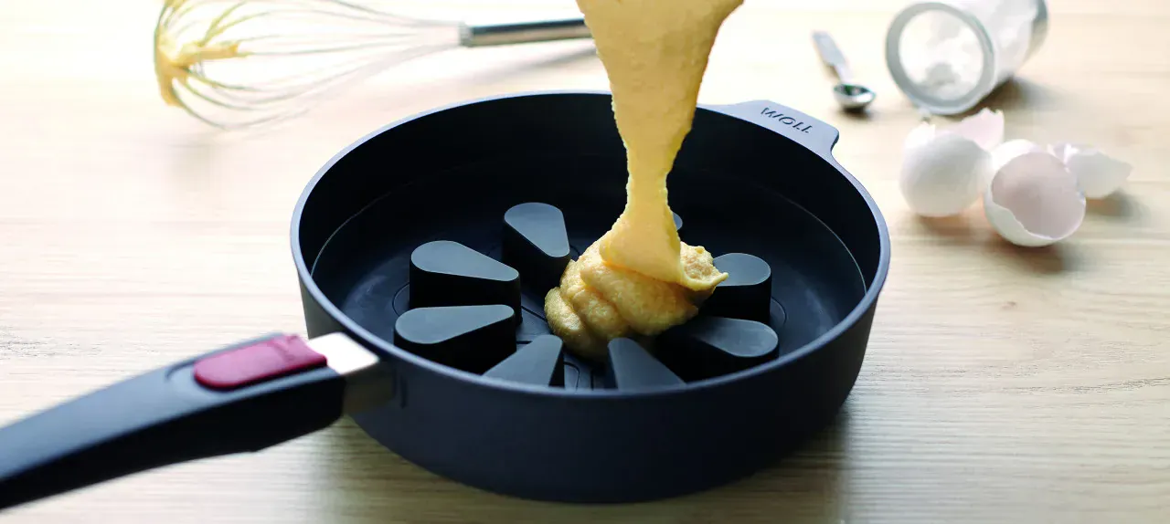 In eine Hochrandpfanne der Serie „Diamond Lite” mit einem Silikon-Backeinsatz der Serie „Bake it” wird heller Teig gefüllt. Im Hintergrund sind ein Schneebesen, ein kleiner Löffel, ein umgefallenes Glas mit Inhalt sowie Eierschalen zu sehen.