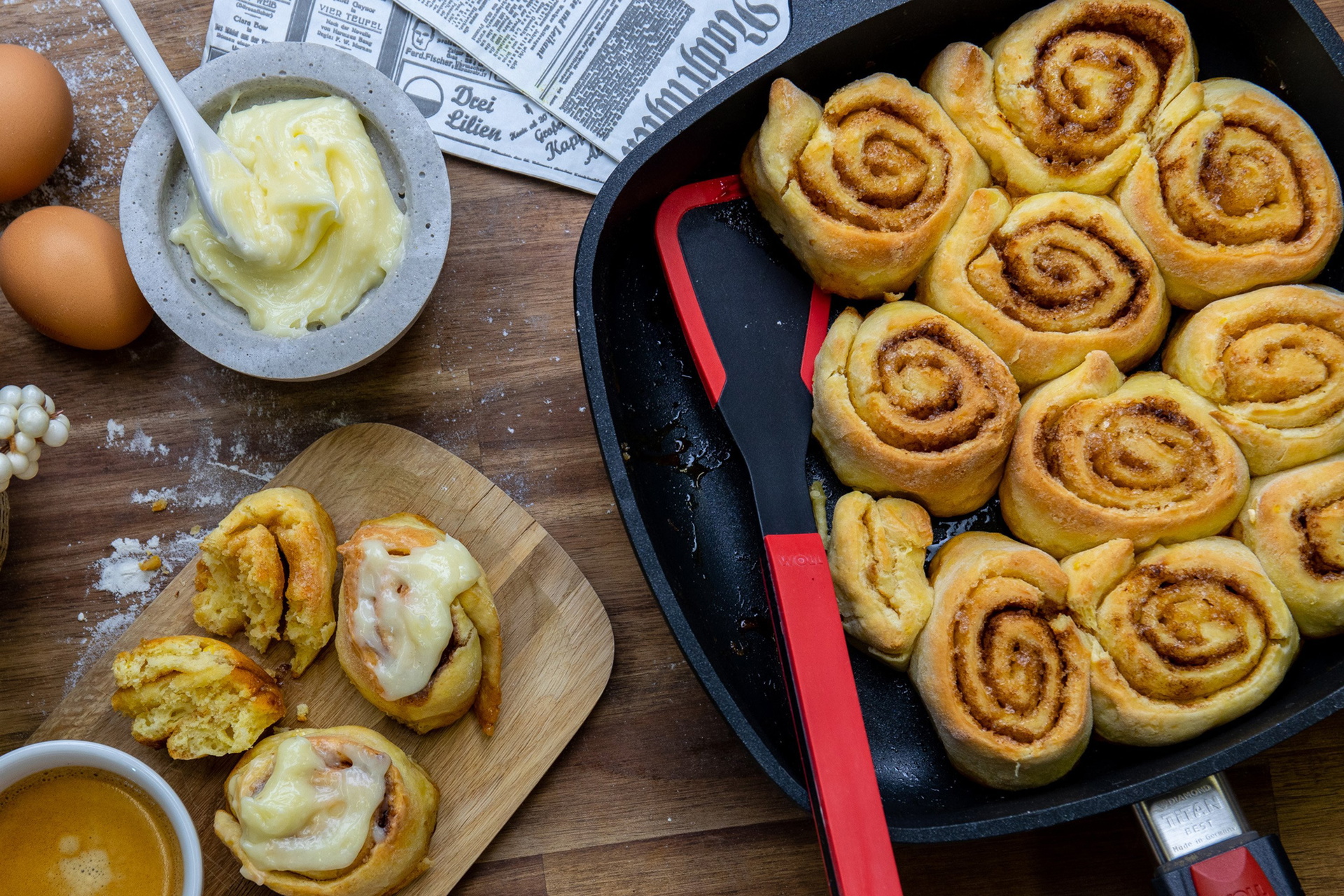 Die Zimtschnecken wurden in einer viereckigen Titan-Best-Pfanne mit abnehmbarem Stiel gebacken. Links daneben liegen entnommene Zimtschnecken auf einem Brett, die mit Topping bestrichen sind. Drumherum liegen Eier und weiteres Topping in einer Schüssel.