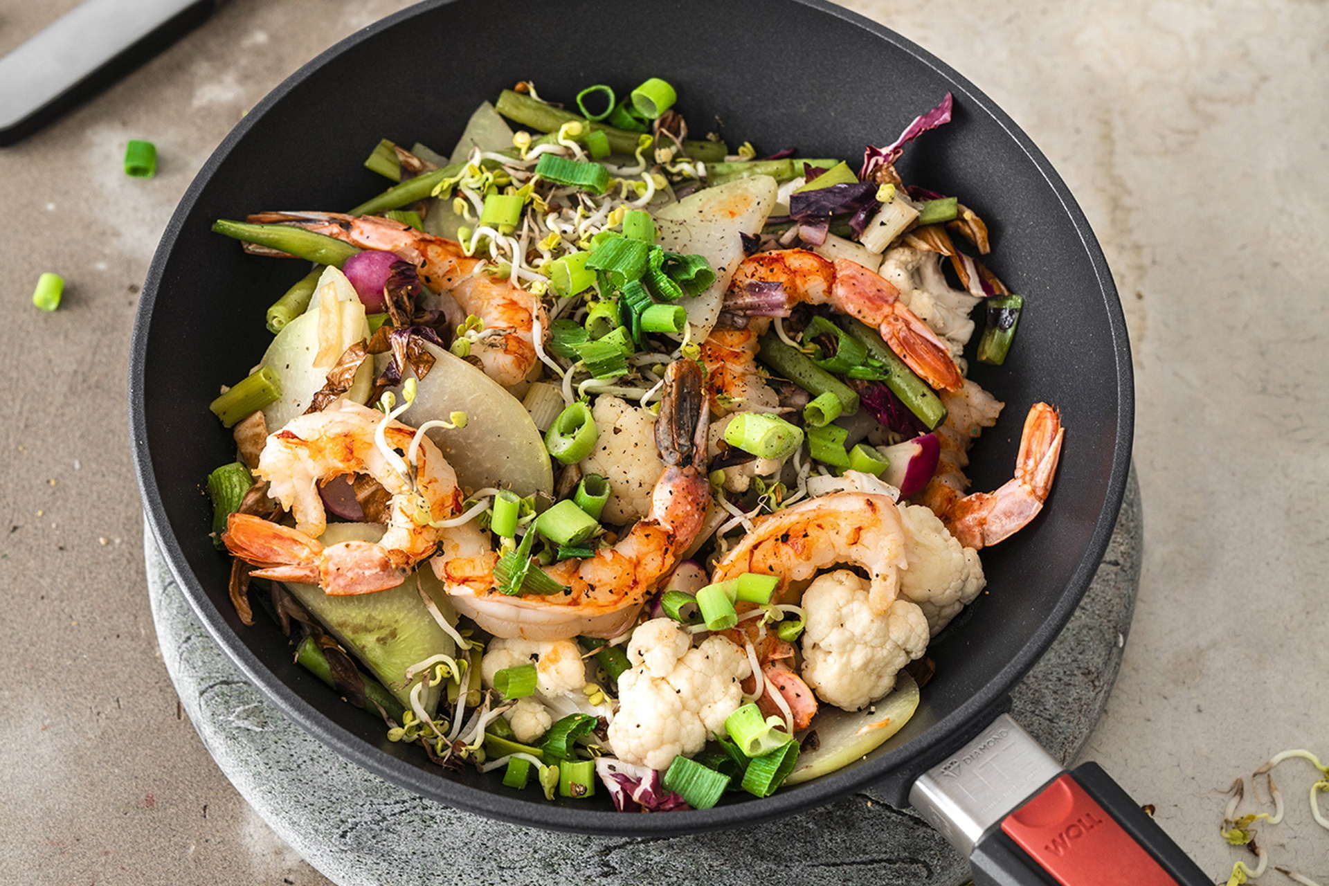 In einer Wok- und Rührpfanne der Serie Diamond Lite befindet sich buntes Sommergemüse mit Garnelen. Dahinter steht eine Schüssel mit Frühlingslauch und weiteren Zutaten.