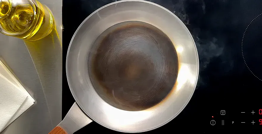 Auf dem Herd steht eine rauchende Carbon-Steel-Pfanne im Einbrennprozess mit leichter Antihaftbeschichtung. Daneben steht eine Flasche mit neutralem Öl, darunter liegt ein Stück Küchenrolle bereit.