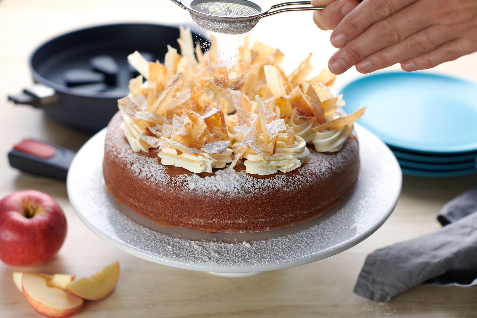 Auf einer Kuchenplatte steht ein frisch gebackener Apfelkuchen, der mit Puderzucker bestäubt wird. Im Hintergrund ist eine runde Diamond-Lite-Pfanne mit abgenommenem Stiel zu sehen. In dieser befindet sich ein Silikon-Backeinsatz. Drumherum stehen noch Te