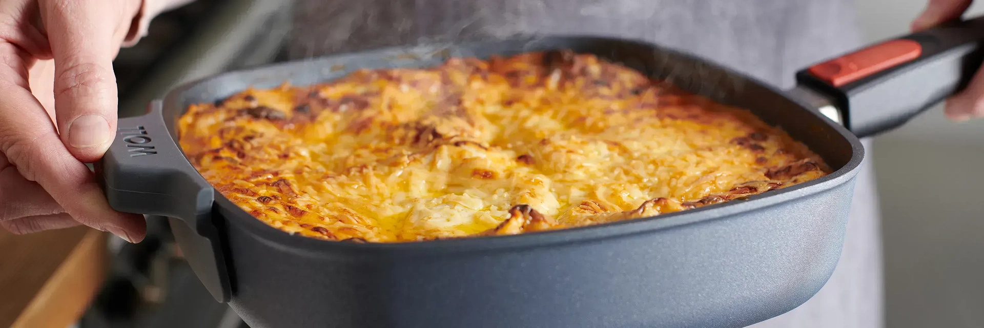 Eine heiße Diamond-Lite-Viereckpfanne mit überbackenem, dampfendem Gericht, frisch aus dem Backofen, wird mit beiden Händen gehalten. Ein aufgesetzter Silikongriff an der Pfanne schützt vor der Hitze.