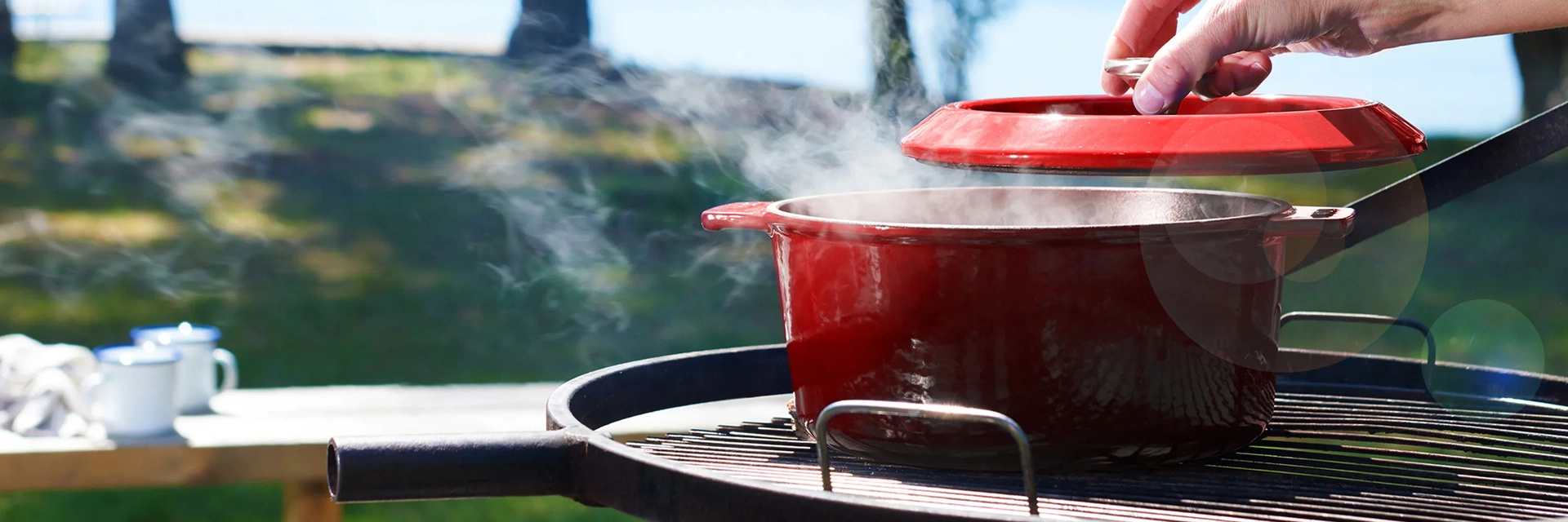 Auf dem Grill steht ein heißer runder Eisenguss-Topf der Serie Iron, in dem sich ein dampfendes Gericht befindet. Der Deckel wurde leicht mit einer Hand angehoben. Im Hintergrund sieht man eine Wiese und Bäume, sowie Tassen auf einem Holzbalken.