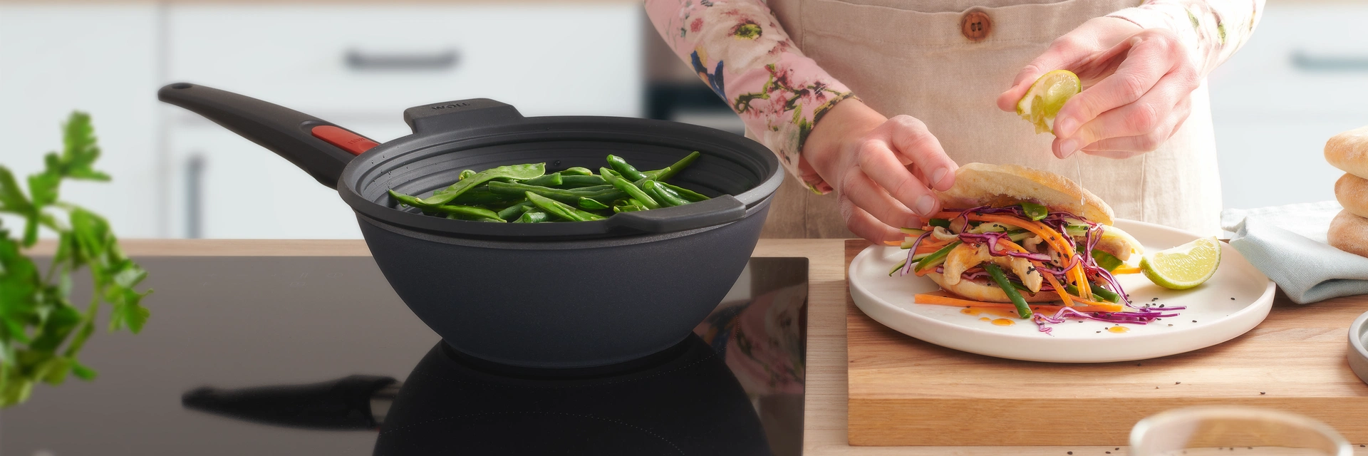 In dem runden Wok der Serie „Diamond Lite” wird mithilfe des Silikoneinsatzes „Steam it” Gemüse gedämpft. Daneben steht ein Teller mit frischem Essen, auf das etwas Limettensaft ausgepresst wird.