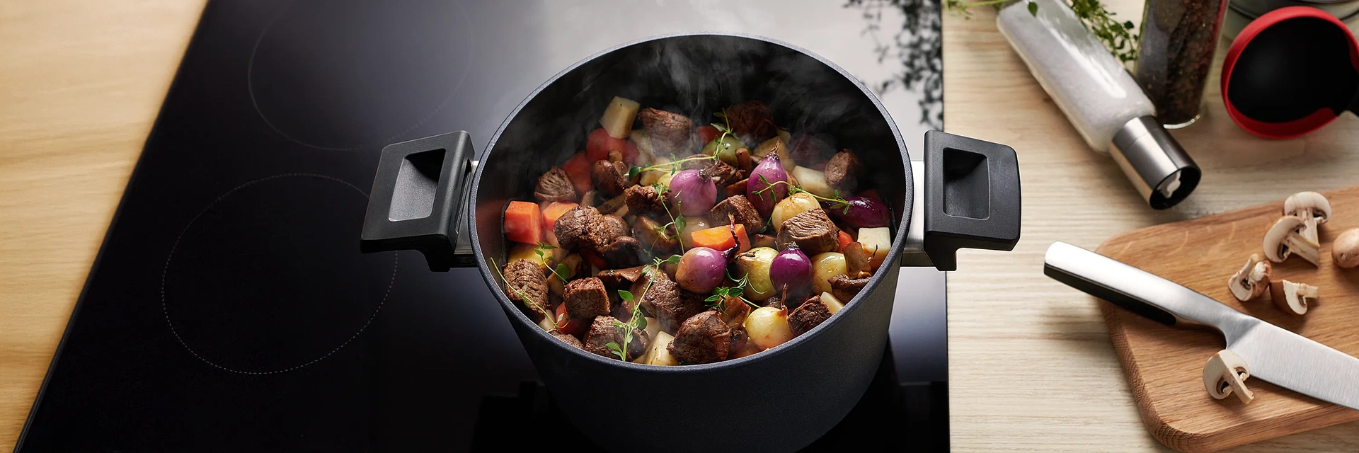 Topf mit Rindergulasch auf einem Herd. Rechts daneben ein Edelstahlmesser und Champignons auf einem Holzbrett, darüber Salz- und Pfefferstreuer.