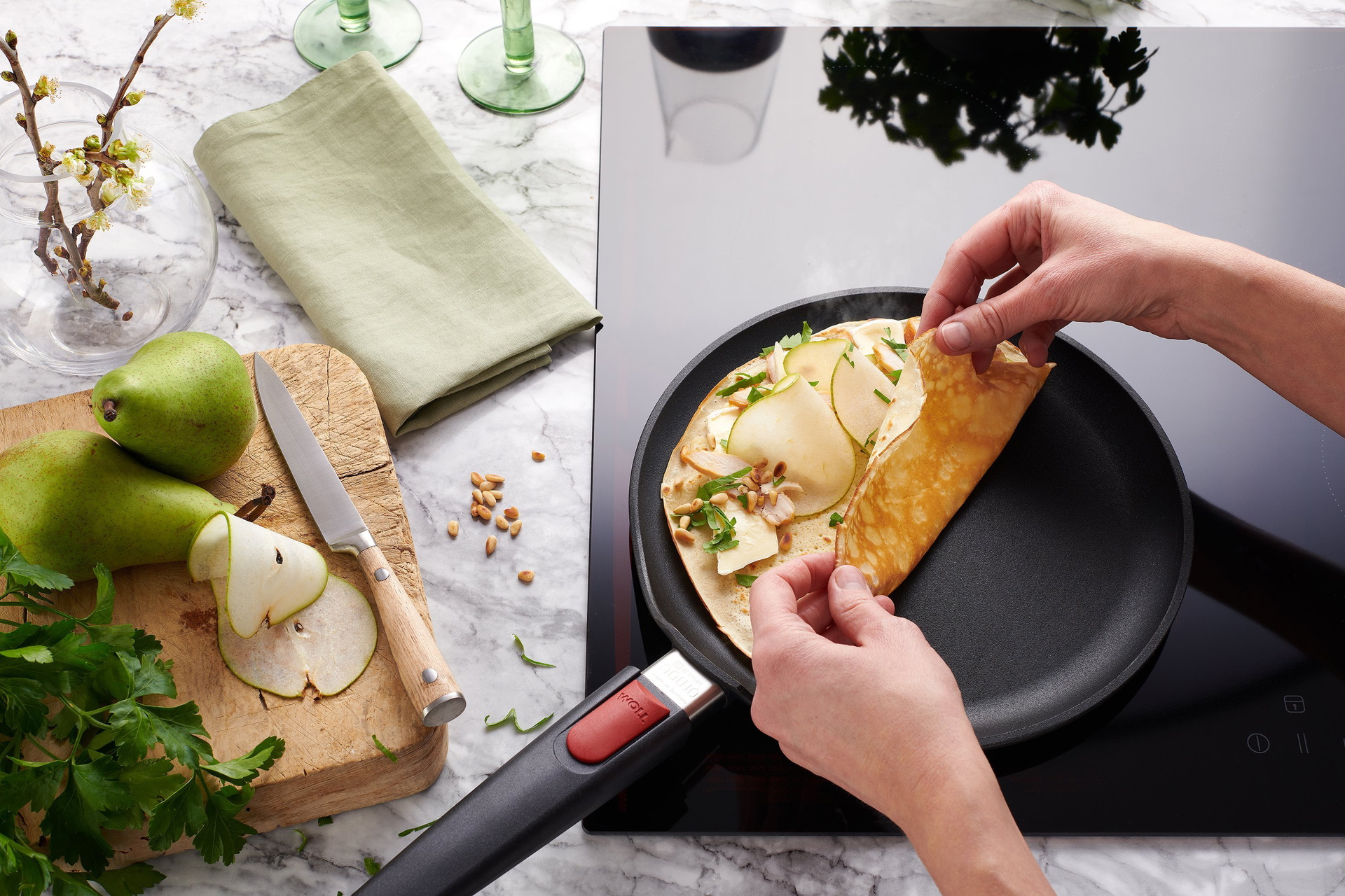 In einer heißen, runden Titan Best Crêpepfanne mit abnehmbarem Stiel wird ein Crêpe mit Brie und Huhn zubereitet. Auf einem Brett daneben liegen zwei ganze Birnen und ein Messer. Darüber befinden sich eine Stoffserviette und zwei Gläser.