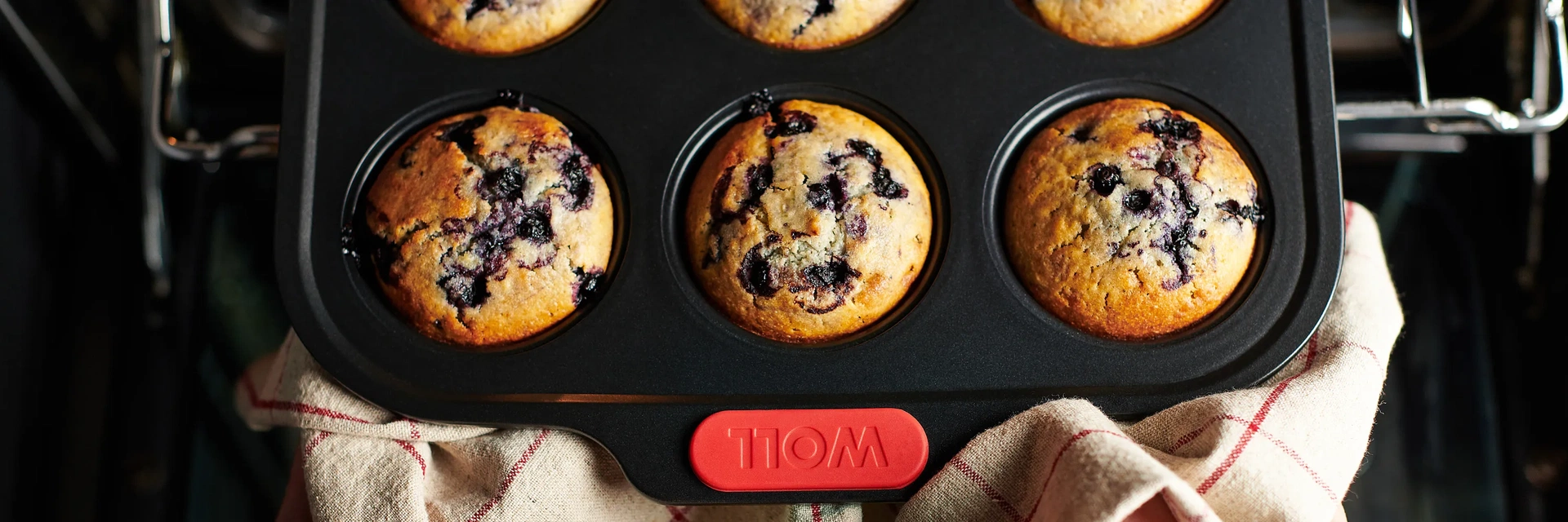 Eine heiße rechteckige Muffinform der Serie Let's Bake! enthält frisch gebackene Blaubeermuffins. Sie wird mit einem Küchentuch aus dem Backofen genommen.