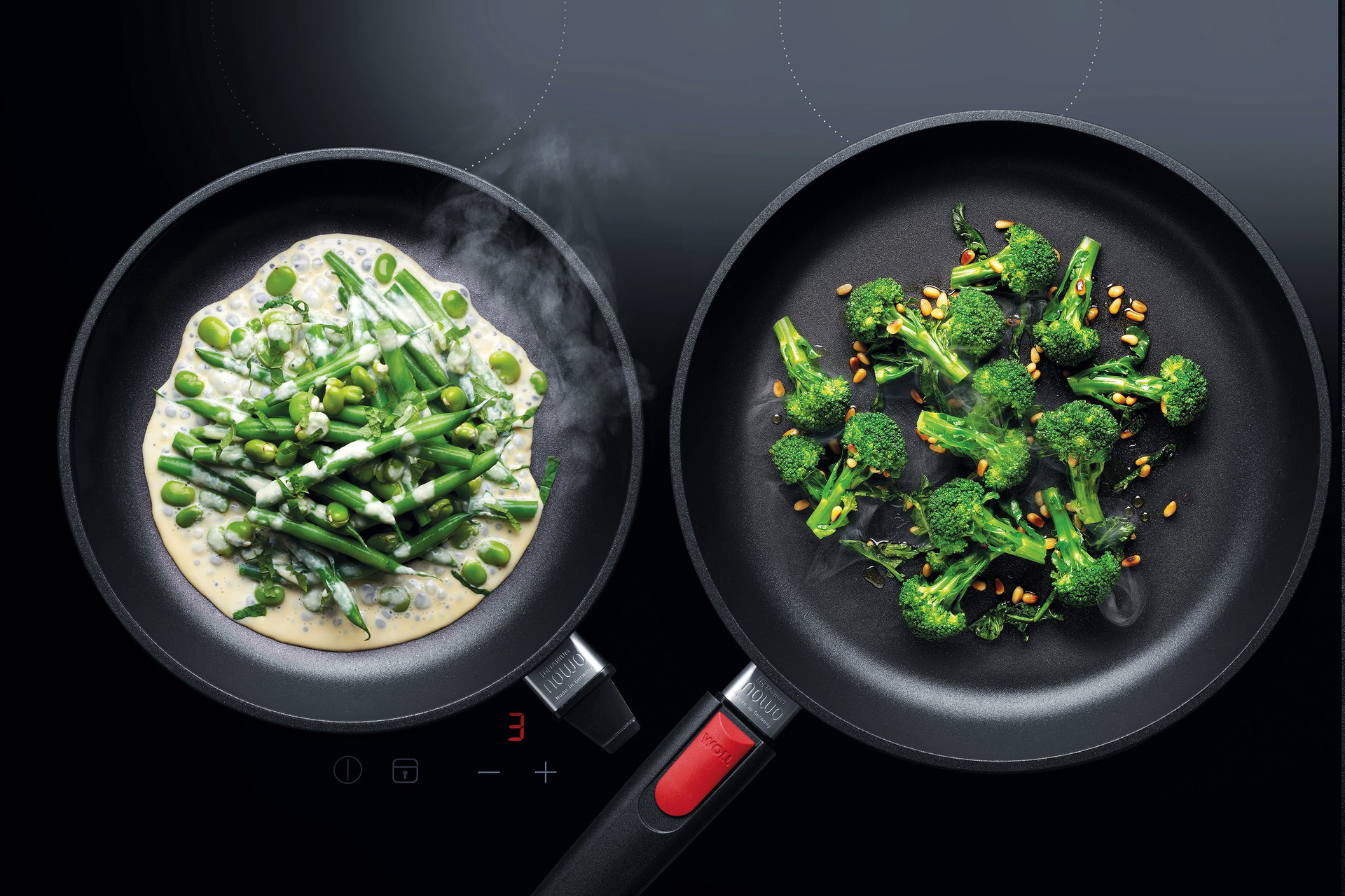 Auf dem Herd stehen zwei heiße, runde Nowo Pfannen. In der linken Pfanne befindet sich Saubohnen und Brechbohnen mit Knoblauchsauce. Um sie sicher neben der rechten Pfanne platzieren zu können, wurde der Stiel entfernt. In der rechten Pfanne, die noch mit