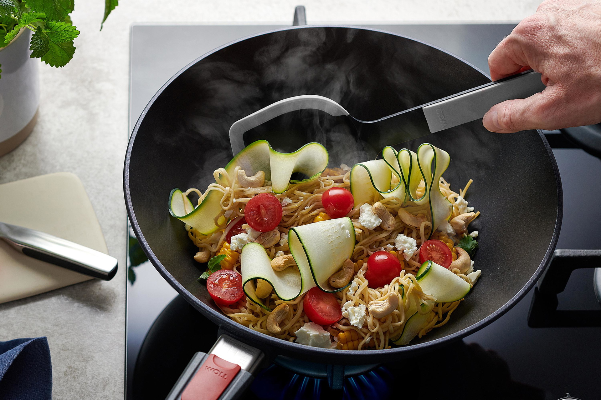In einer runden Diamond Lite Wok- und Rührpfanne wird ein Nudelgericht zubereitet, das mit einer grauen Cock it Pfannenschaufel umgerührt wird. 