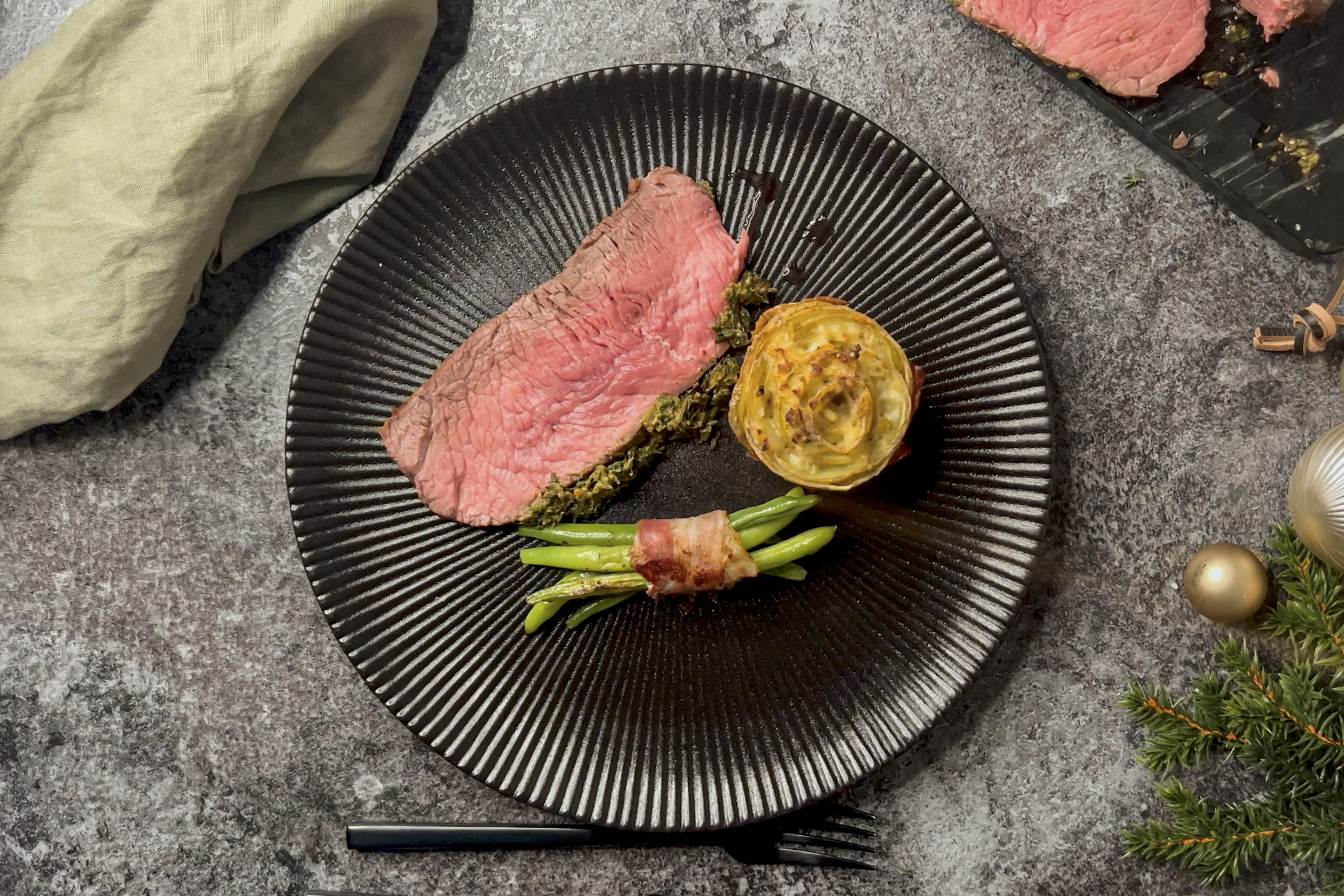 Auf einem Teller liegt Roastbeef mit Kräuterkruste, daneben wurden eine Rosenkartoffel und Bohnen im Speckmantel platziert. Auf dem Tisch liegen eine Stoffserviette, ein Schneidebrett mit weiterem Roastbeef sowie weihnachtliche Dekoration.