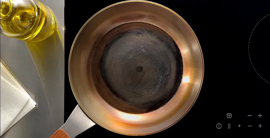 Auf dem Herd steht eine Carbon-Steel-Pfanne mit stark eingebrannter Antihaftbeschichtung. Daneben stehen eine Flasche mit neutralem Öl und darunter liegt ein Stück Küchenrolle bereit.