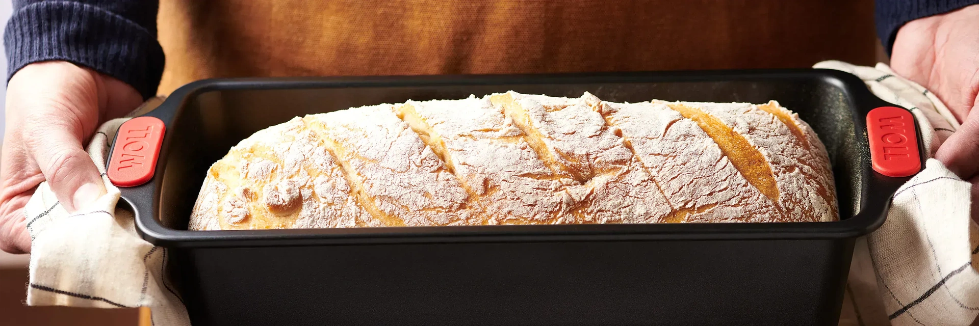 Eine heiße, rechteckige Kastenform der Serie Let's Bake! wird mit frischem Brot mit beiden Händen an den Griffen mithilfe eines Küchentuchs gehalten.