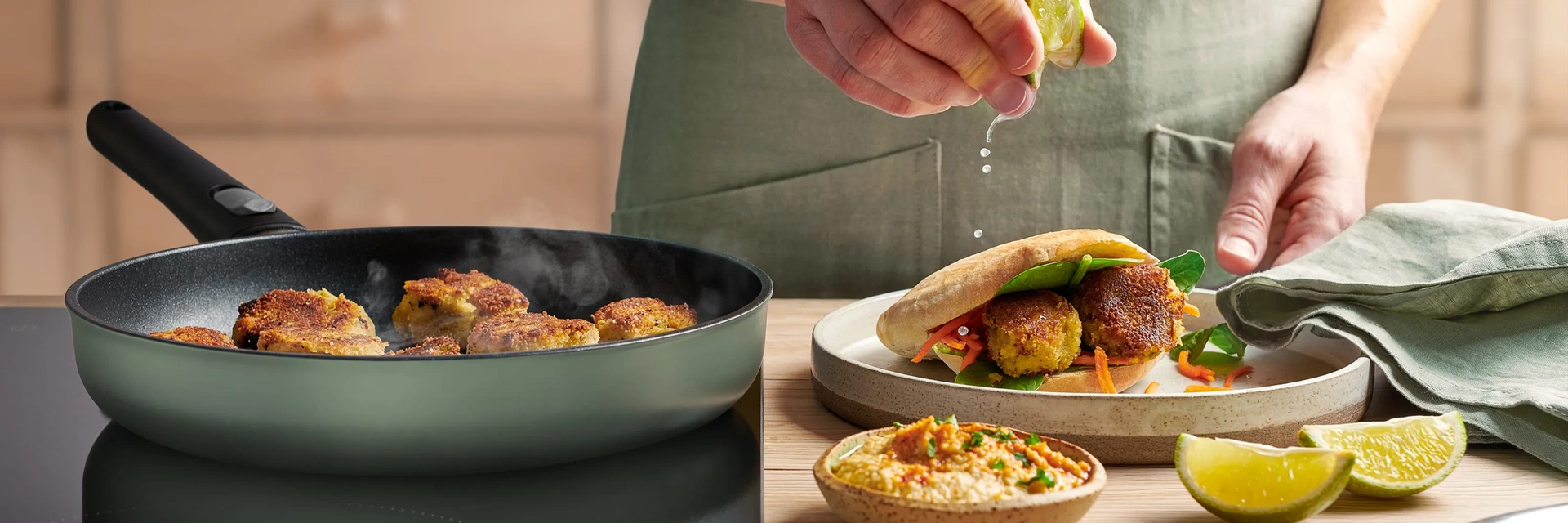 In einer runden, heißen Ecolour Pfanne werden frische Falafel angebraten. Auf dem Teller daneben liegt ein fertiges Fladenbrot gefüllt mit Falafel und eine Schüssel Hummus steht vor dem Teller.