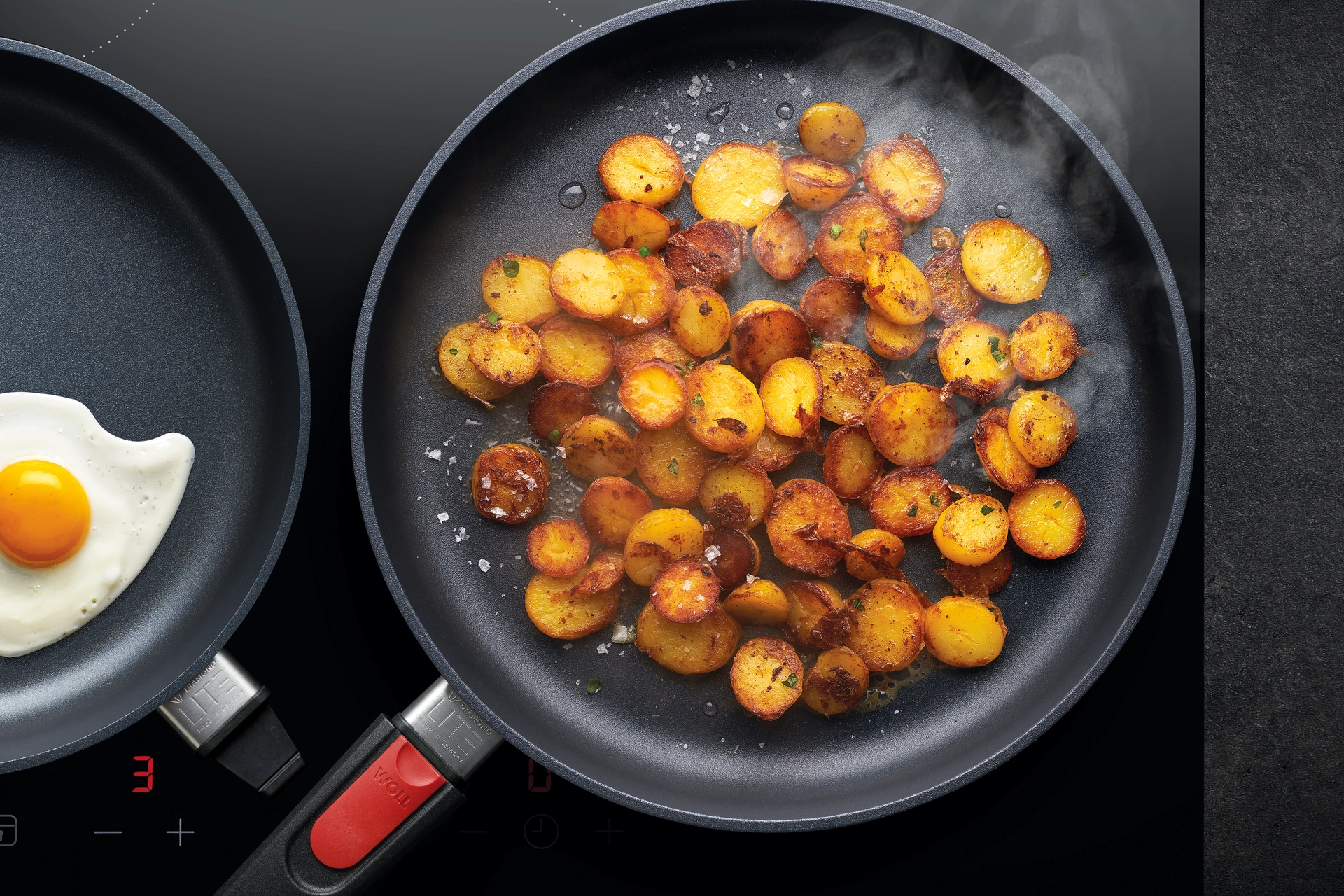 In einer runden Diamond Lite Pfanne werden Bratkartoffeln angebraten, während in einer weiteren Pfanne Spiegeleier zubereitet werden. 