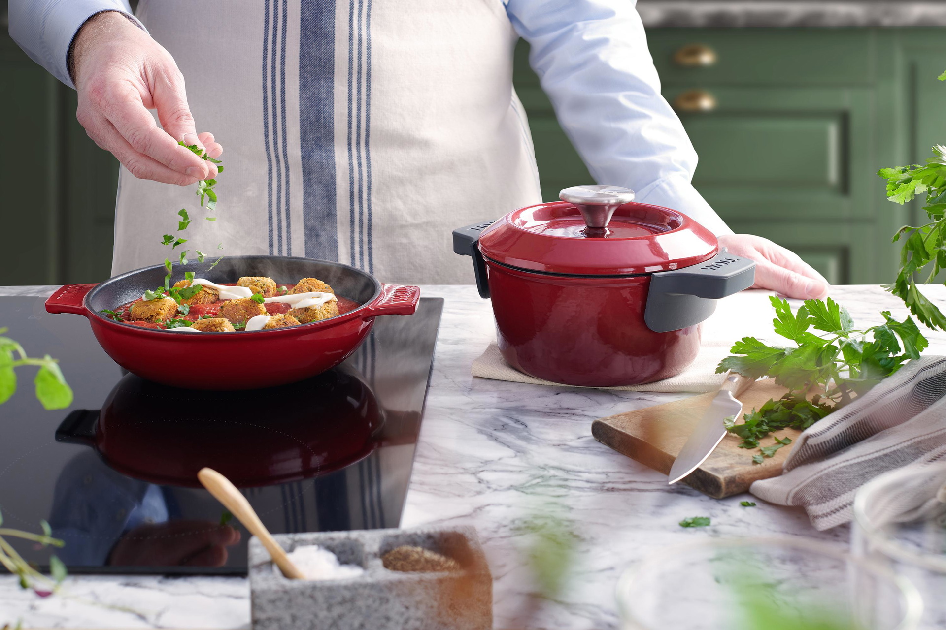 5520CI-010_116CI-010_Mood Auf dem Herd steht eine rote, runde Iron Servierpfanne, in der sich ein dampfendes Gericht befindet. Daneben steht ein Iron Topf mit Deckel.