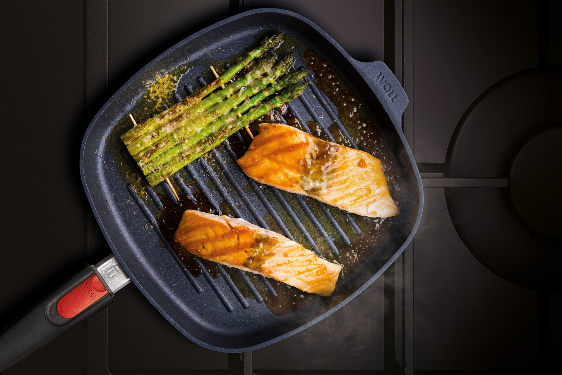 Auf dem Herd steht eine viereckige, heiße Diamond-Lite-Grillpfanne mit abnehmbarem Stiel. Darin befindet sich frisch zubereiteter, dampfender Honig-Senf-Lachs mit grünem Spargel.
