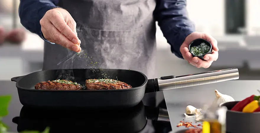Auf dem Herd steht die rechteckige, heiße Logic-Grillpfanne mit abnehmbarem Edelstahlstiel, in der saftig angebratenes Fleisch dampft. Es ist mit Kräutersalz bestreut. Daneben liegen frische Knoblauchzehen auf der Arbeitsplatte und in einem Behälter stehe