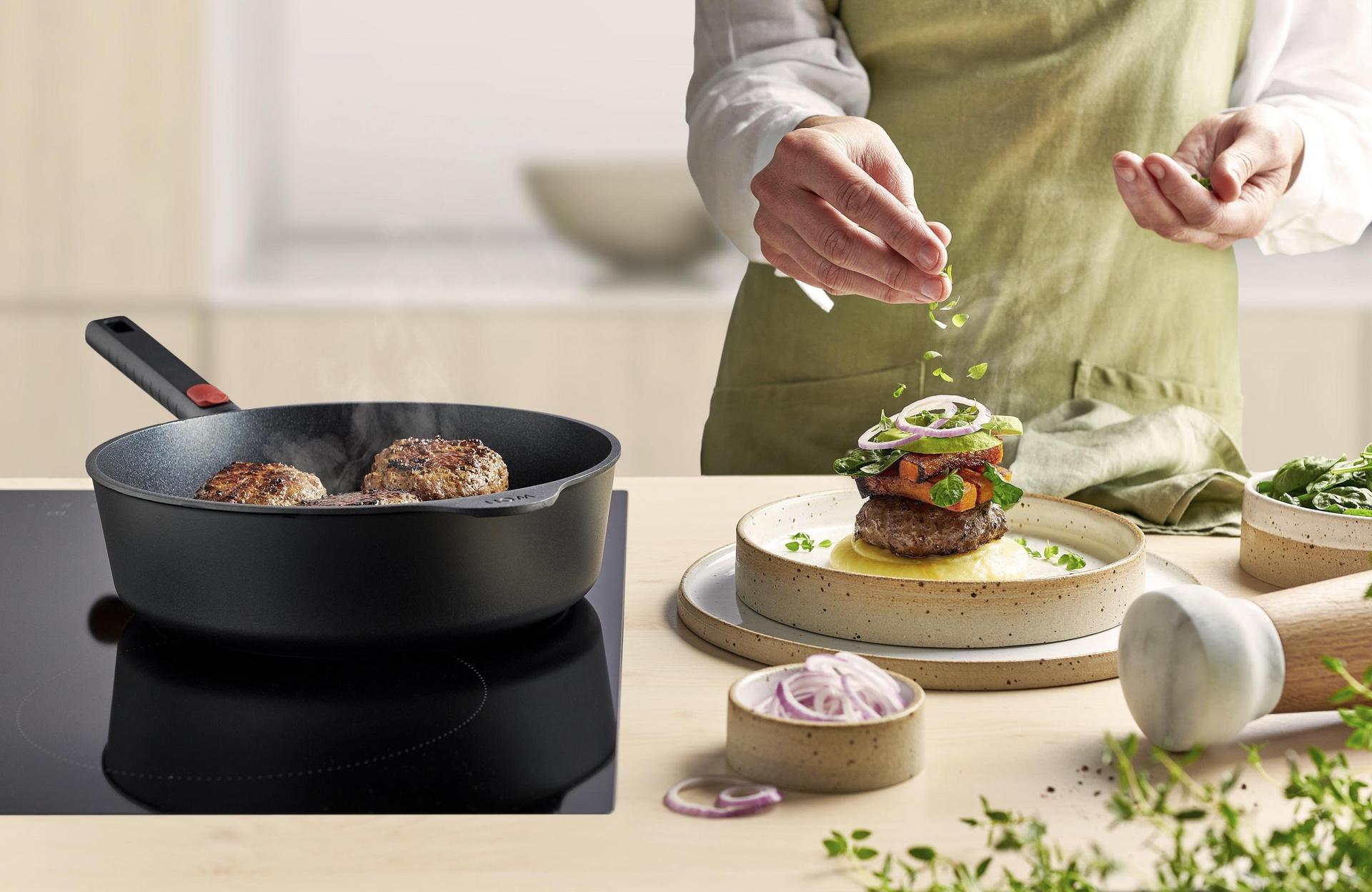 Auf dem Herd steht eine runde, heiße Eco-Logic-QXR Pfanne mit abnehmbarem Stiel, in der frische Pilze angebraten werden. Auf einem Brett steht eine Schüssel mit frisch angemachten Bandnudeln mit Pilzen, die mit Parmesan garniert werden. Davor stehen ein r