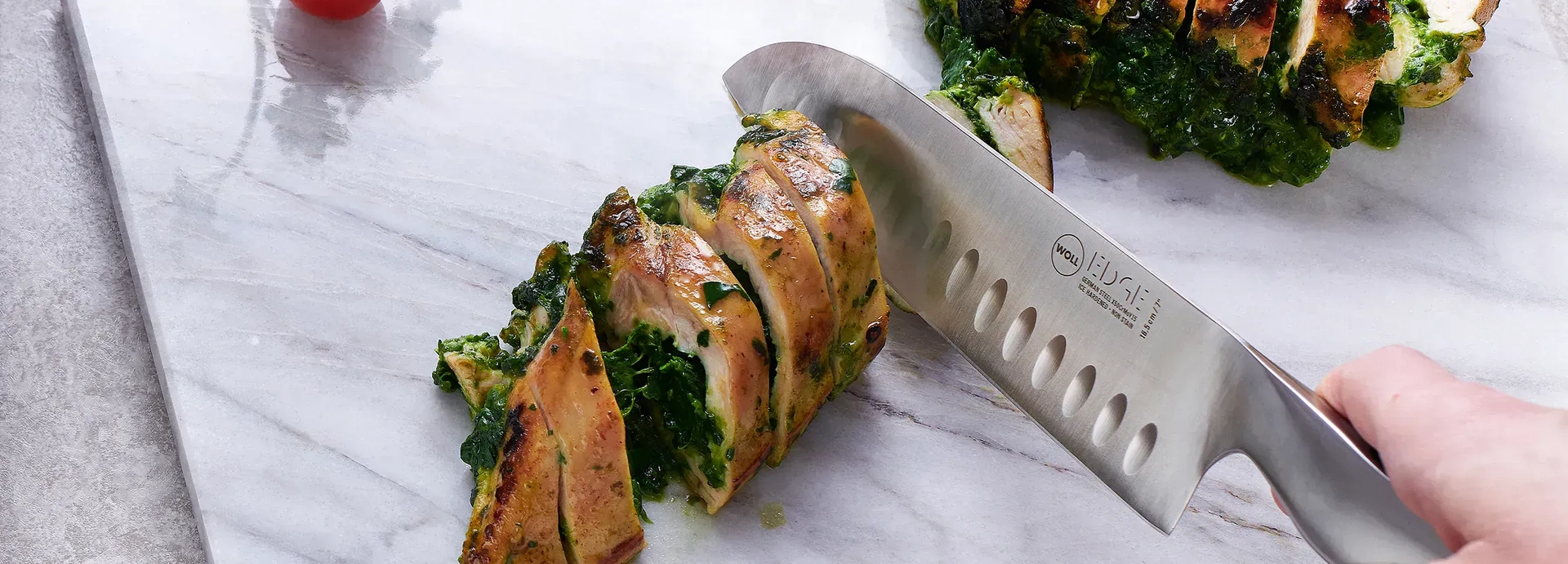 Mit einem Messer der Serie „Edge“ wird eine mit Spinat gefüllte Fleischrolle auf dem Schneidebrett in Portionsgrößen geschnitten. Daneben liegen Cherrytomaten und eine Handvoll Spinat.