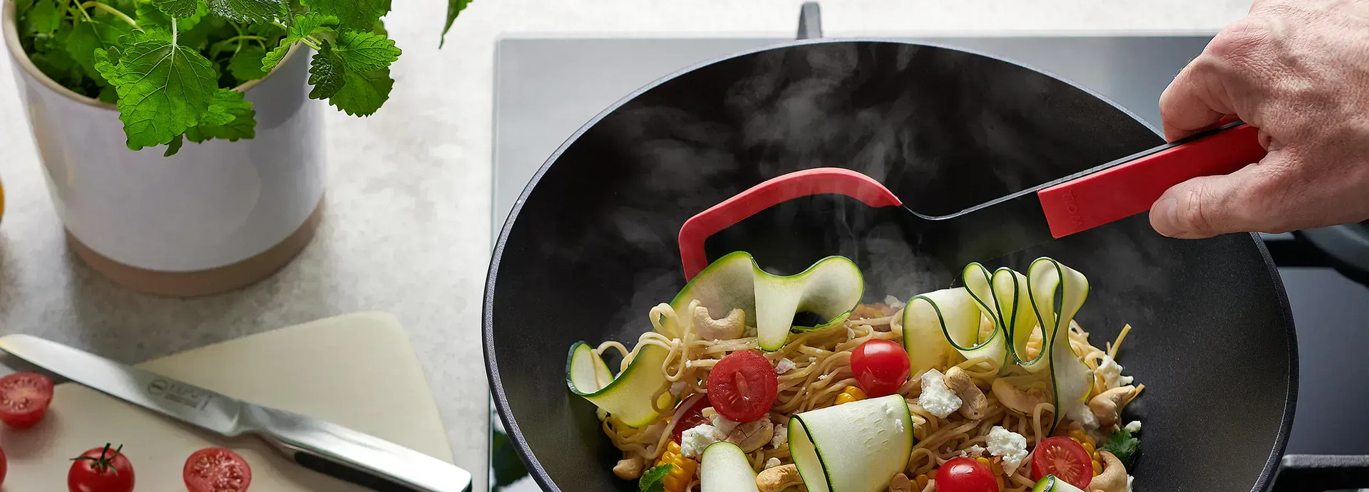 Auf dem Herd steht eine runde Diamond-Lite-Wokpfanne, in der ein dampfendes Nudelgericht zubereitet wird. Links daneben steht ein Topf mit Kräutern, auf dem Schneidebrett liegen ein Messer und halbierte Cherrytomaten.