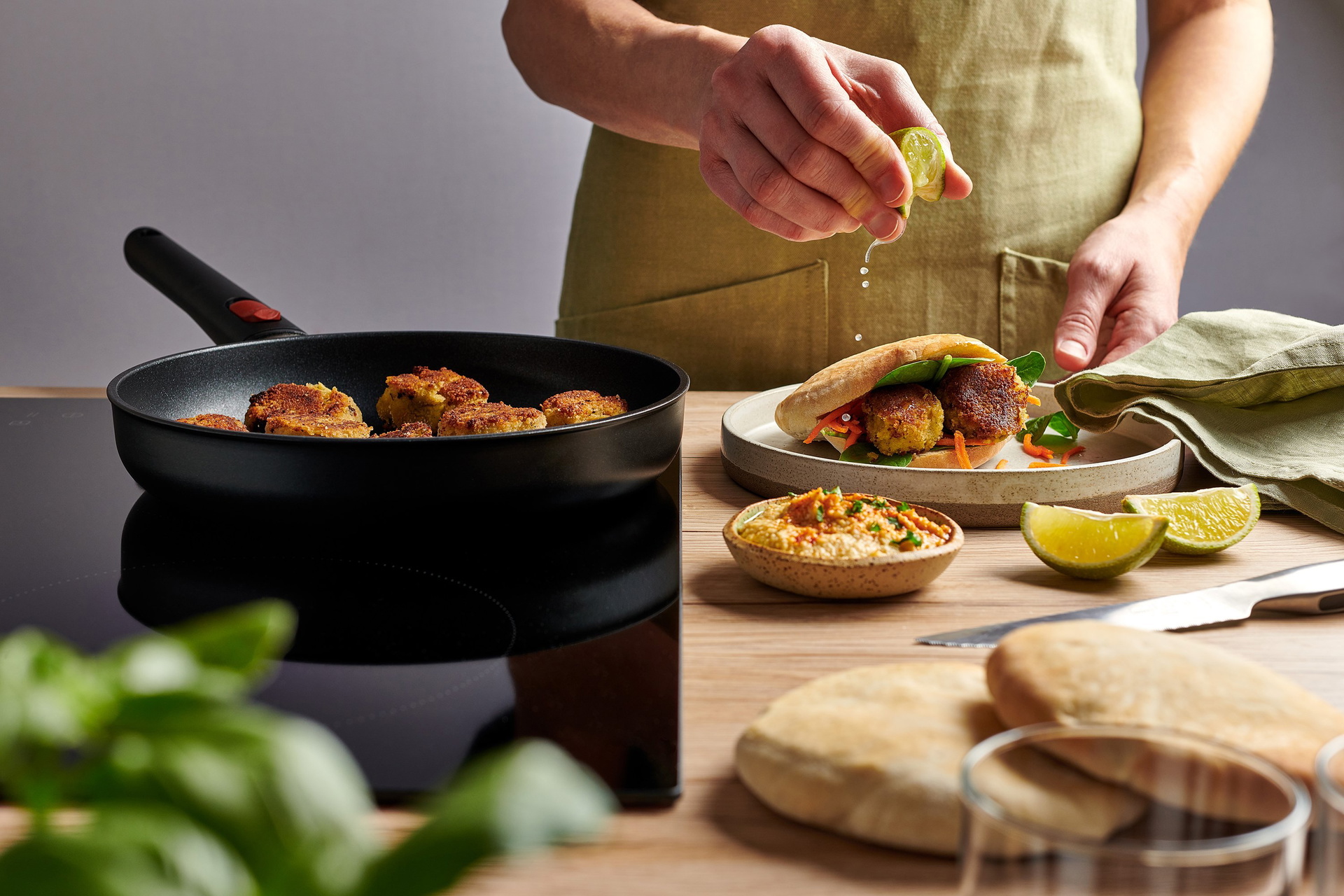 In einer heißen, runden Eco-Lite-QXR-Pfanne werden frische Falafel angebraten. Daneben auf dem Teller liegt ein fertiges Fladenbrot, das mit Falafel gefüllt ist, sowie eine Schüssel Hummus, die mit frischem Limettensaft verfeinert wird.