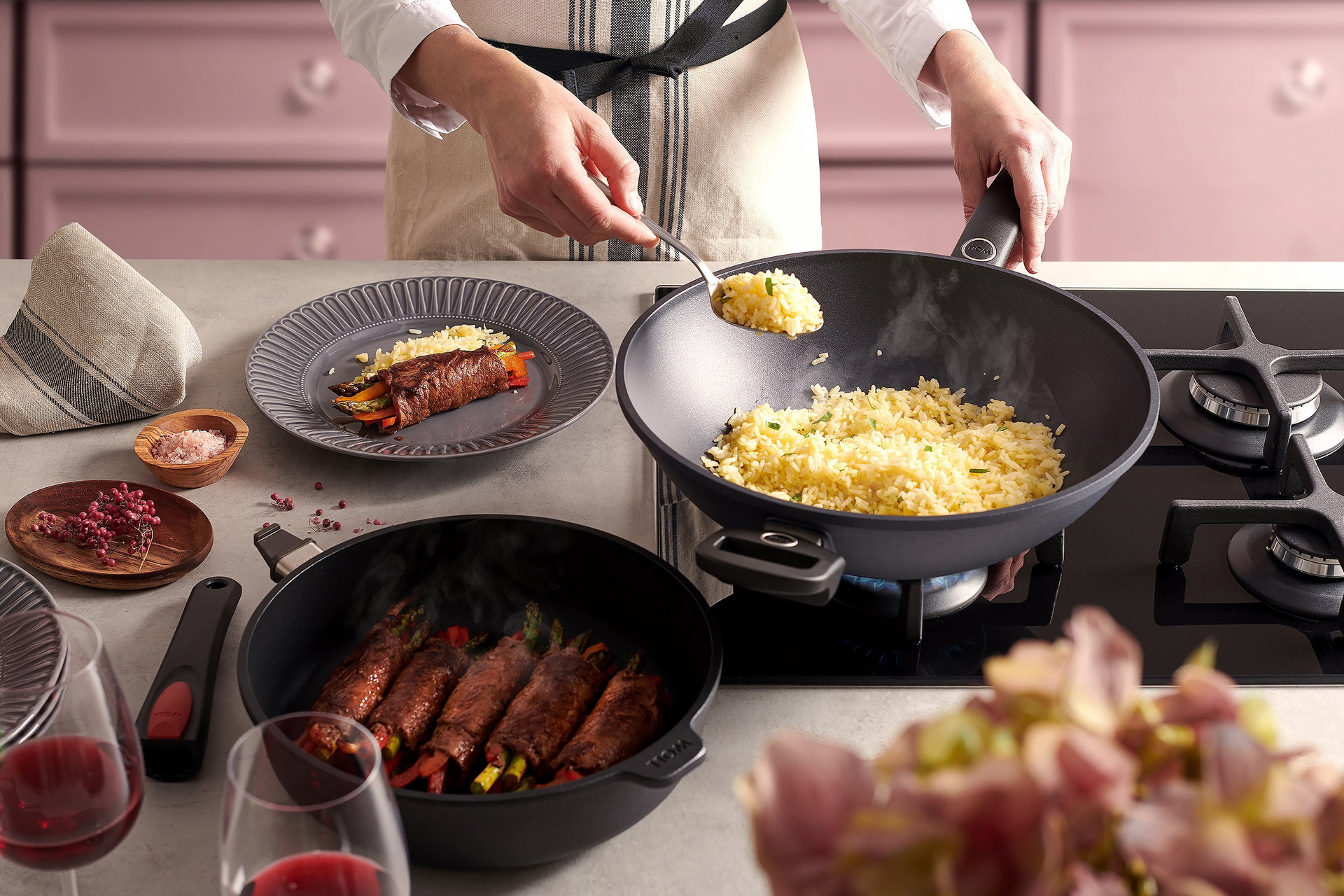 Auf dem Herd steht eine runde, heiße Wok- und Rührpfanne der Serie Titan Best mit festem Stiel, in der Reis zubereitet wird. Auf der Arbeitsplatte daneben steht eine Pfanne der Serie Titan Best mit abgenommenem Stiel, in der sich dampfender Spargel befin