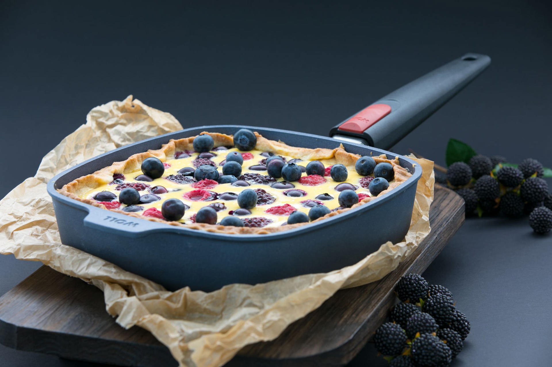 Auf einem Holzbrett steht eine viereckige Diamond-Lite-Pfanne mit abnehmbarem Stiel. In dieser befindet sich eine frisch gebackene Beerentorte. Daneben liegen frische Beeren.