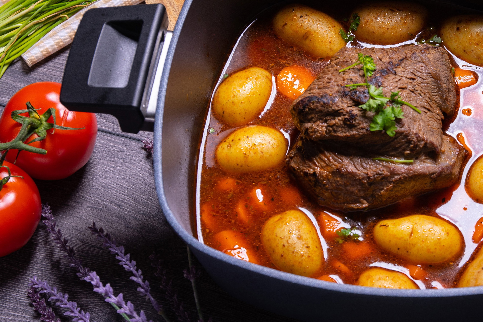 In einem heißen, viereckigen Diamond Active Lite Niederdrucktopf befindet Rinderbraten mit Kartoffeln. Daneben liegen ganze Tomaten und weitere Zutaten.