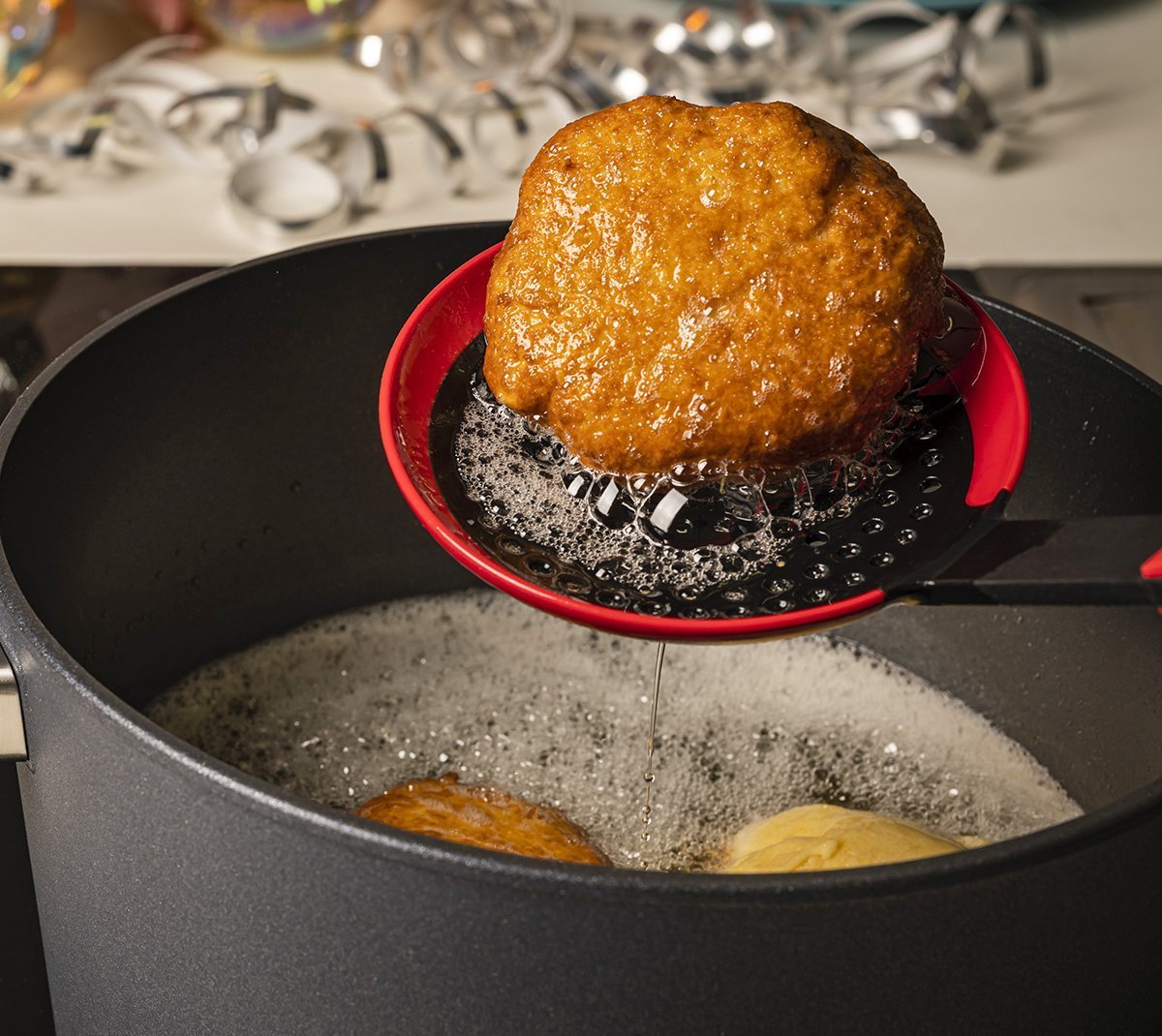 KU006_Mood_1200x1070_RGB Mit einer roten Cock it Schöpfkelle wird ein ausbackener Fastnachtkrapfen aus dem Topf entnommen und über dem Topf abgetropft.