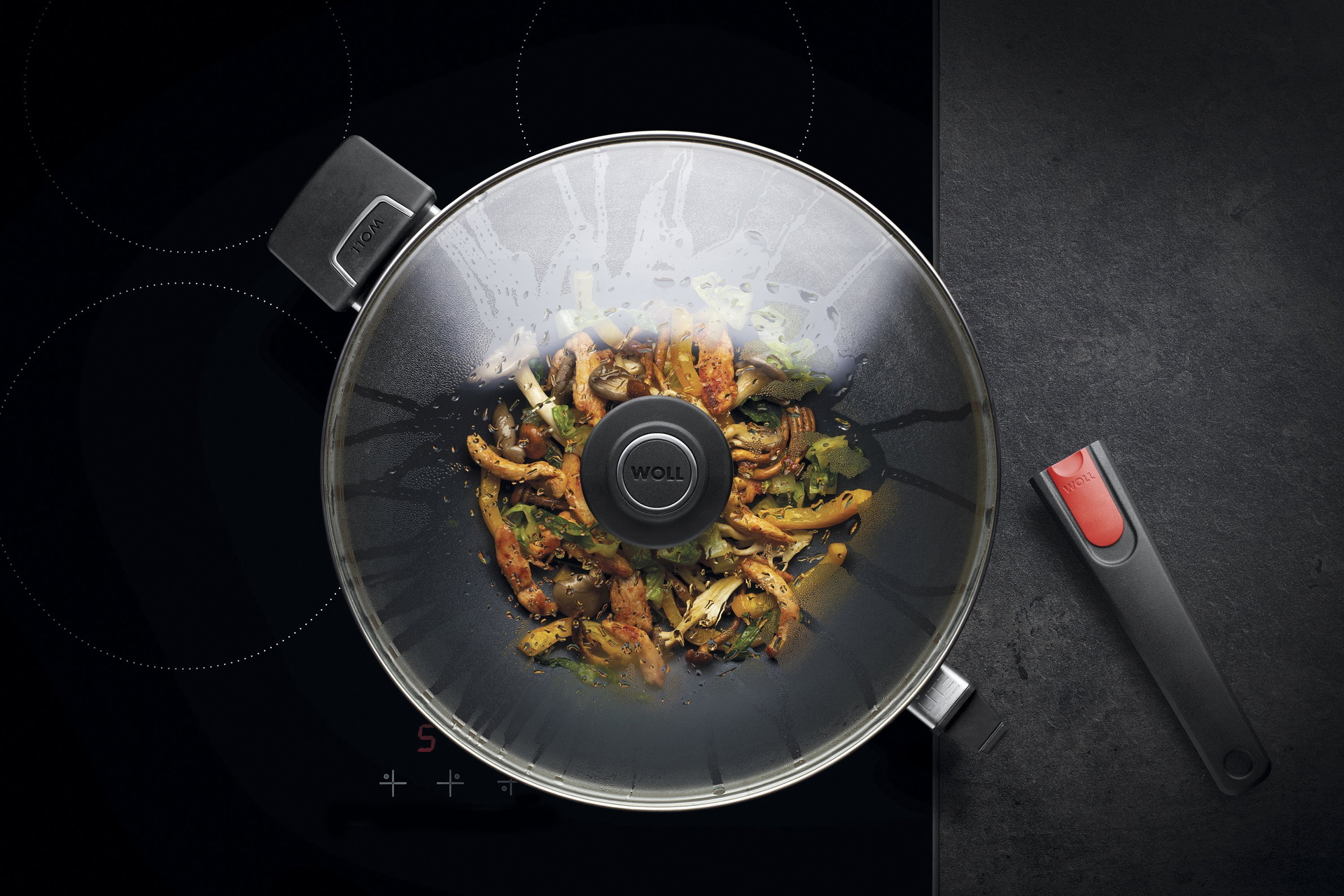 Auf dem Herd steht eine runde Diamond Lite Wok- und Rührpfanne  In dieser befindet sich ein  frisch zubereitetes Gericht. Am Glasdeckel hat sich Kondenswasser gebildet.