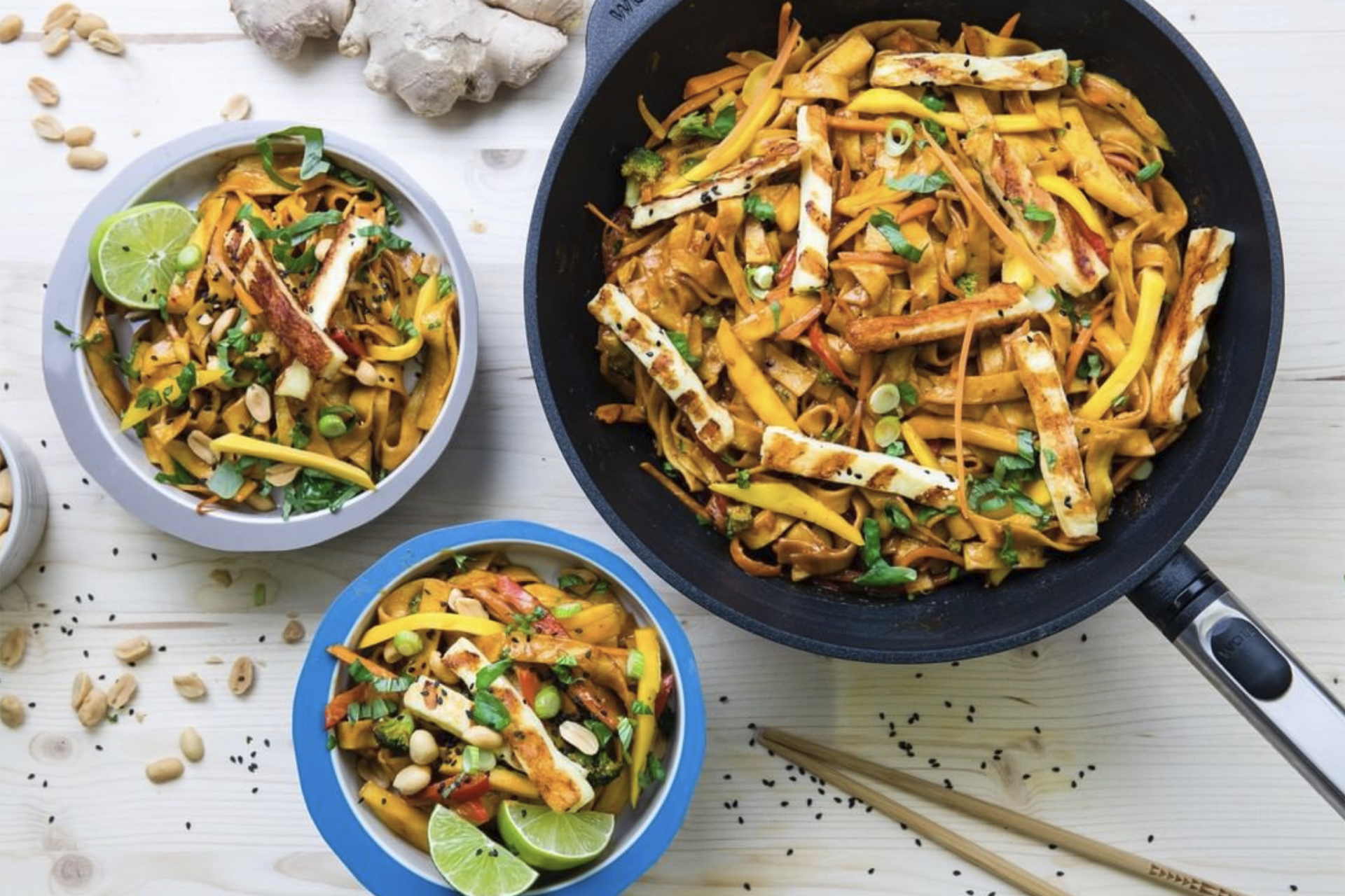 In einer heißen, runden Logic Wok- und -Rührpfanne mit abnehmbarem Stiel befinden sich frisch zubereitete Erdnuss Nudeln mit Halloumi. Daneben stehen Schüsseln mit dem Gericht. Drumherum liegen Erdnüsse und Essstäbchen.